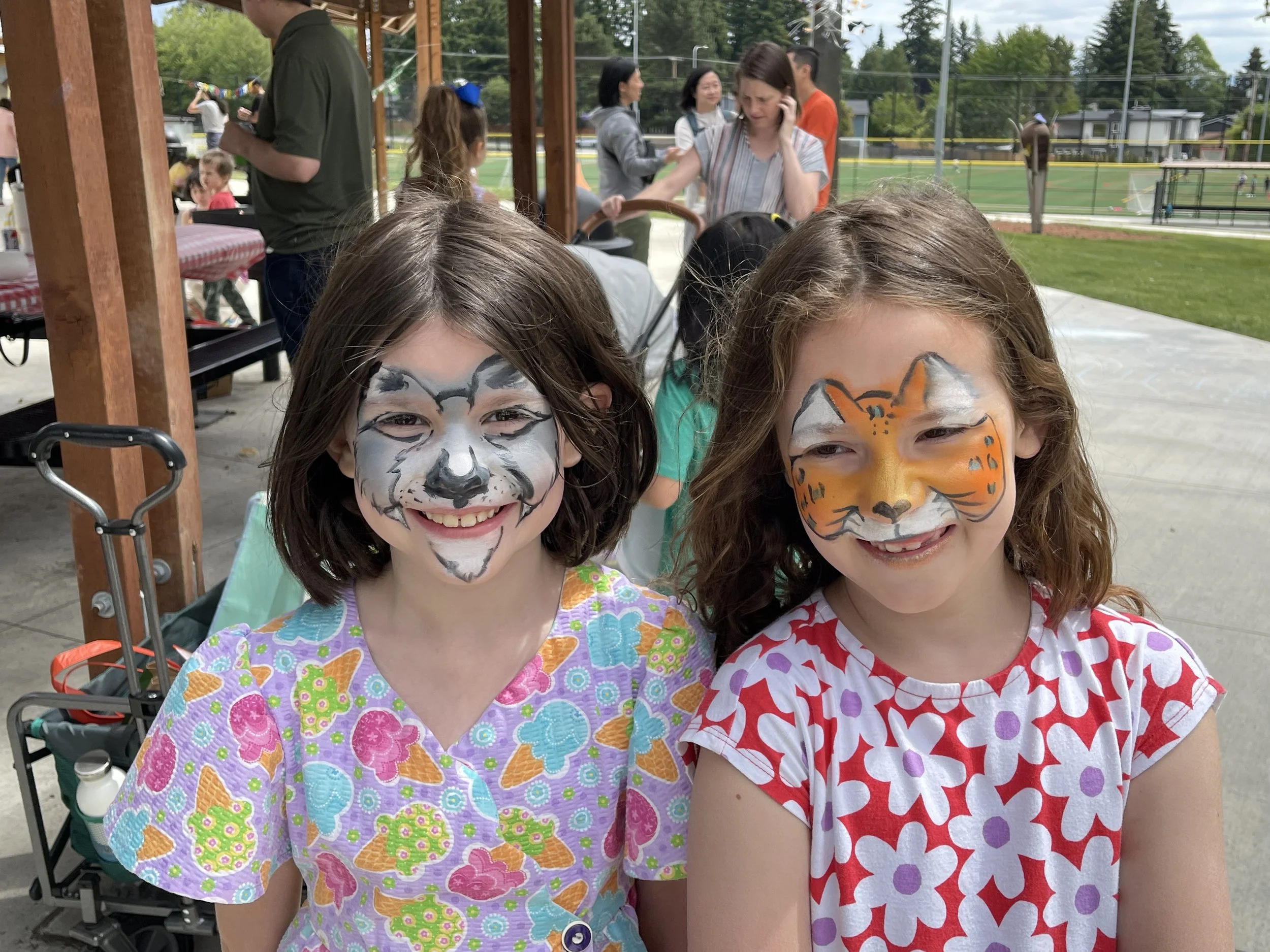 Two smiling girls with cat and tiger face paint pose for a photo at an outdoor gathering.