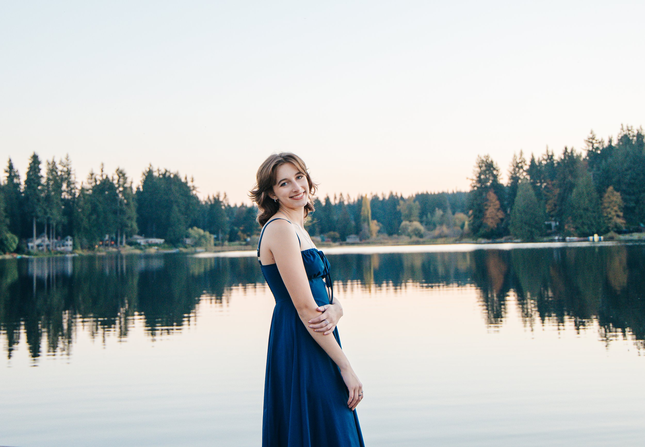 Smiling woman in a blue dress standing beside a calm lake at sunset, with trees reflected in the water.