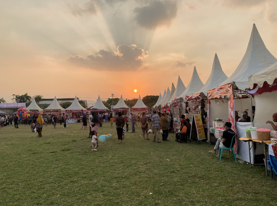 Outdoor festival with white peaked vendor tents, food stalls, and crowds gathering at sunset.