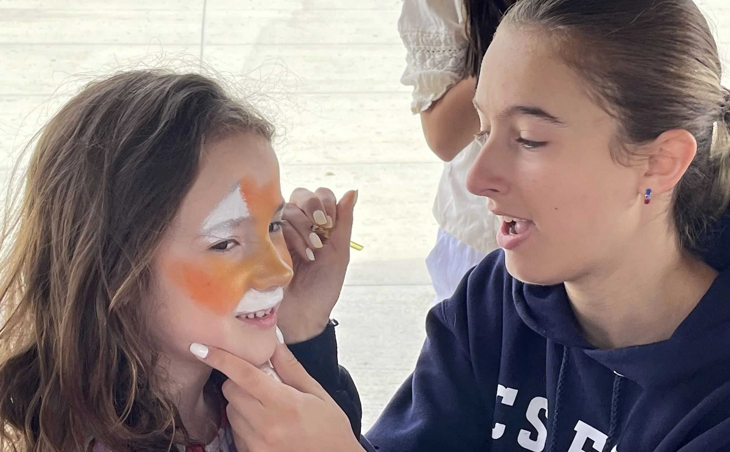 Woman applying white and orange face paint to a smiling child while another person stands behind them