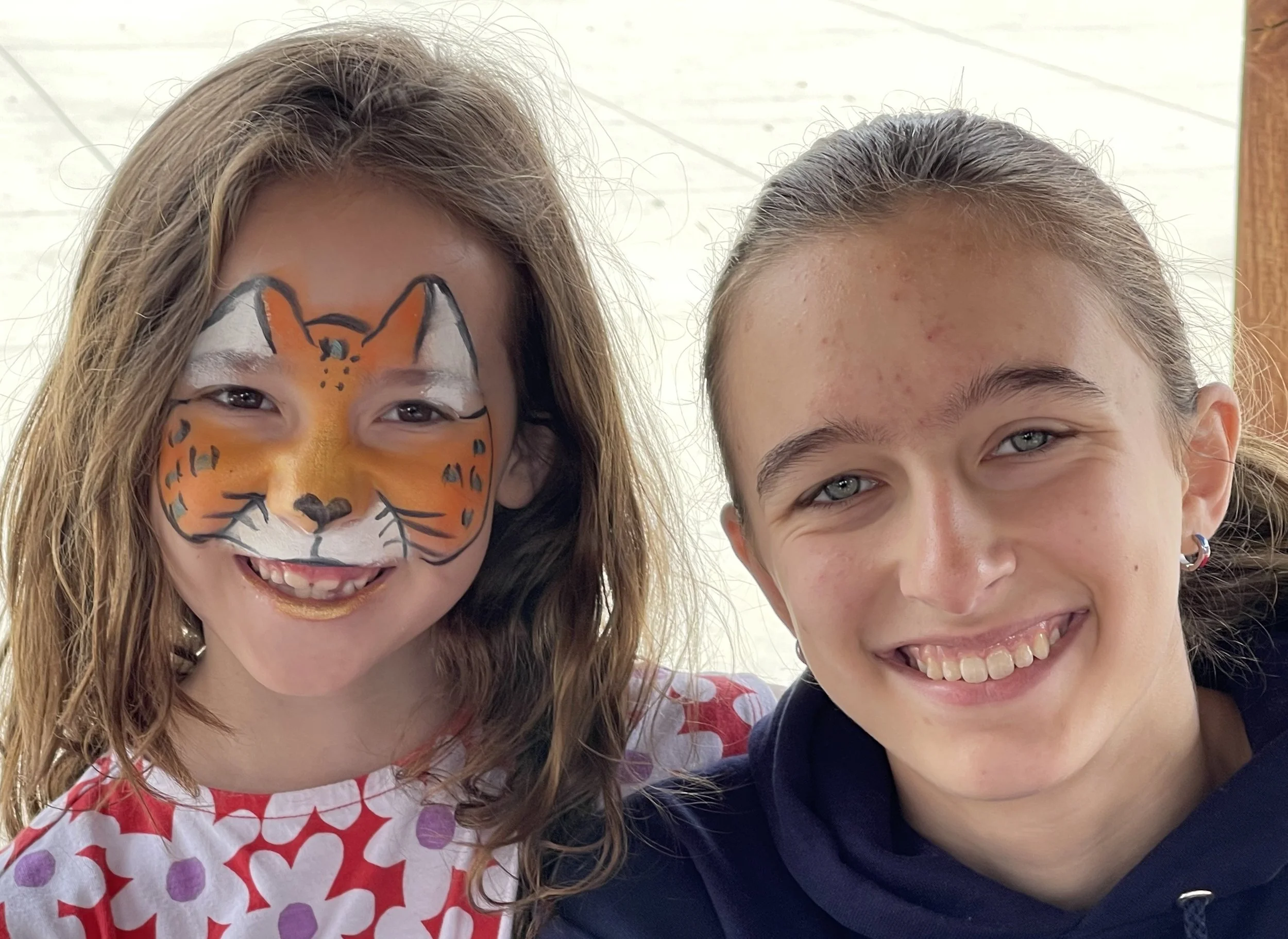 Two smiling girls taking a selfie, one wearing colorful tiger face paint.