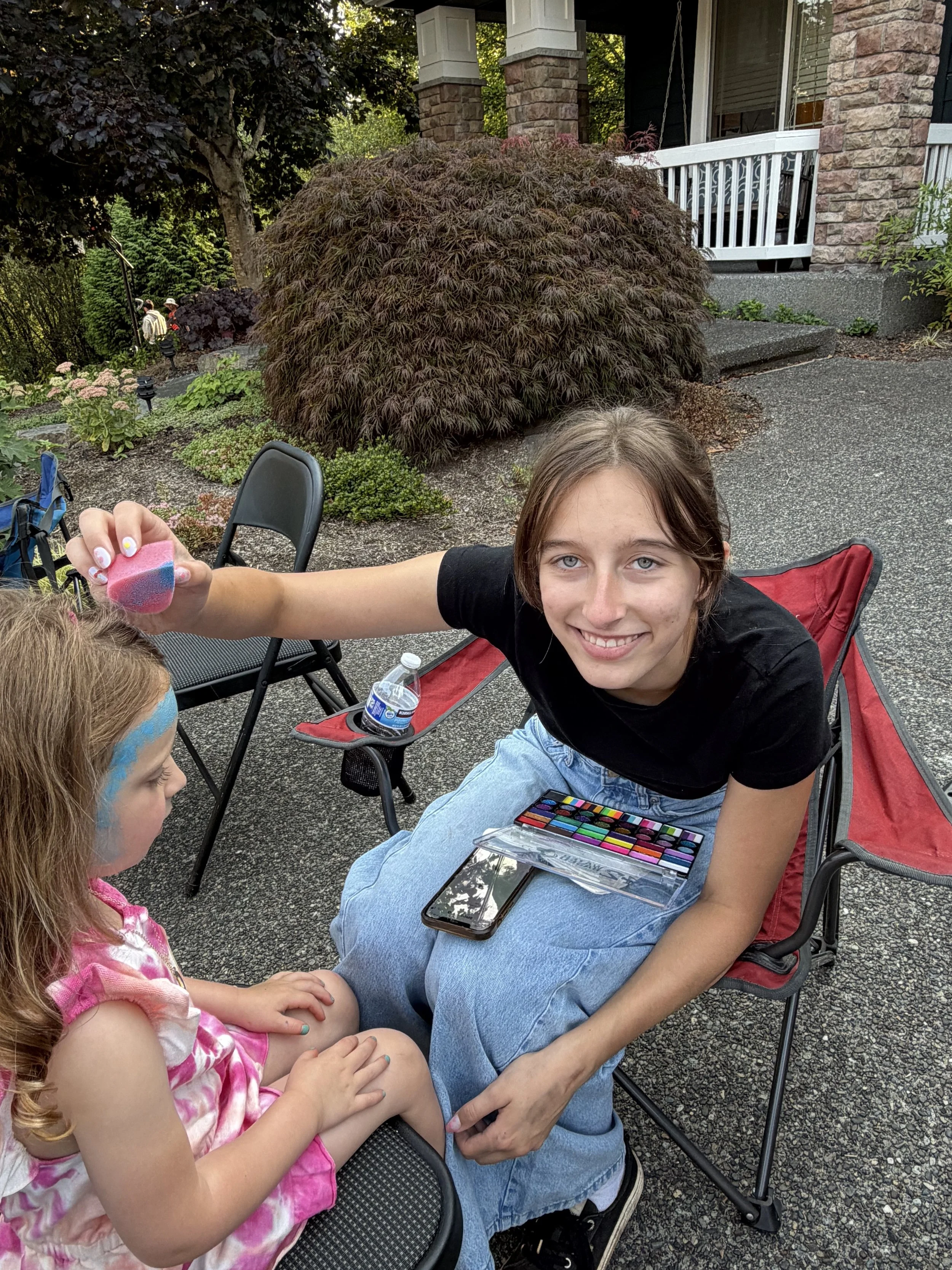 Two girls seated outdoors in chairs, with one holding a colorful cup beside a landscaped garden and stone porch.