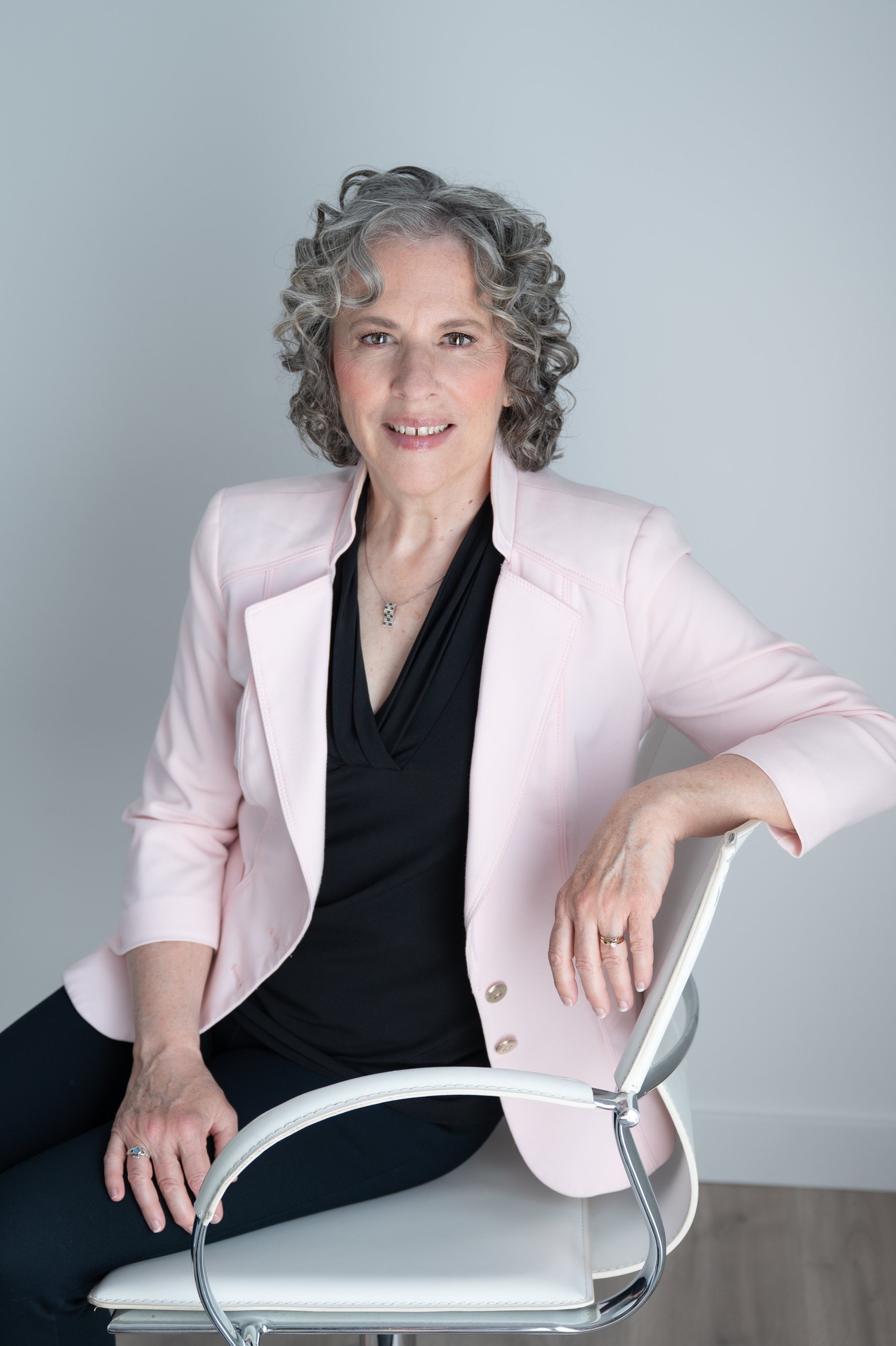Professional portrait of a smiling older woman with curly gray hair wearing a pink blazer and black dress while seated in a white chair