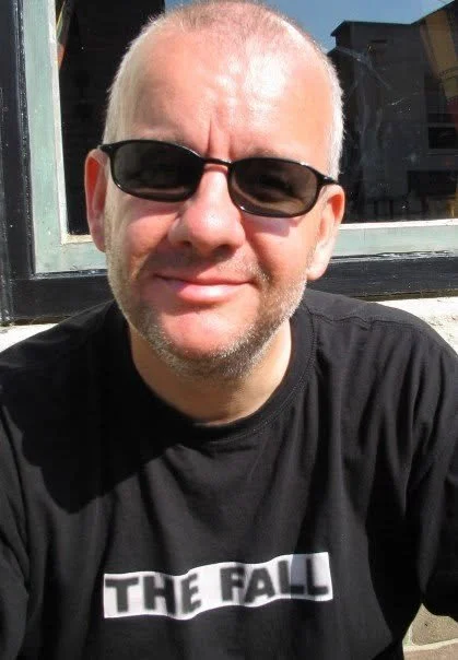 Bald man wearing sunglasses and a black The Fall T-shirt outdoors