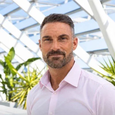 Portrait of a smiling bearded man in a pink shirt standing in a bright glass greenhouse