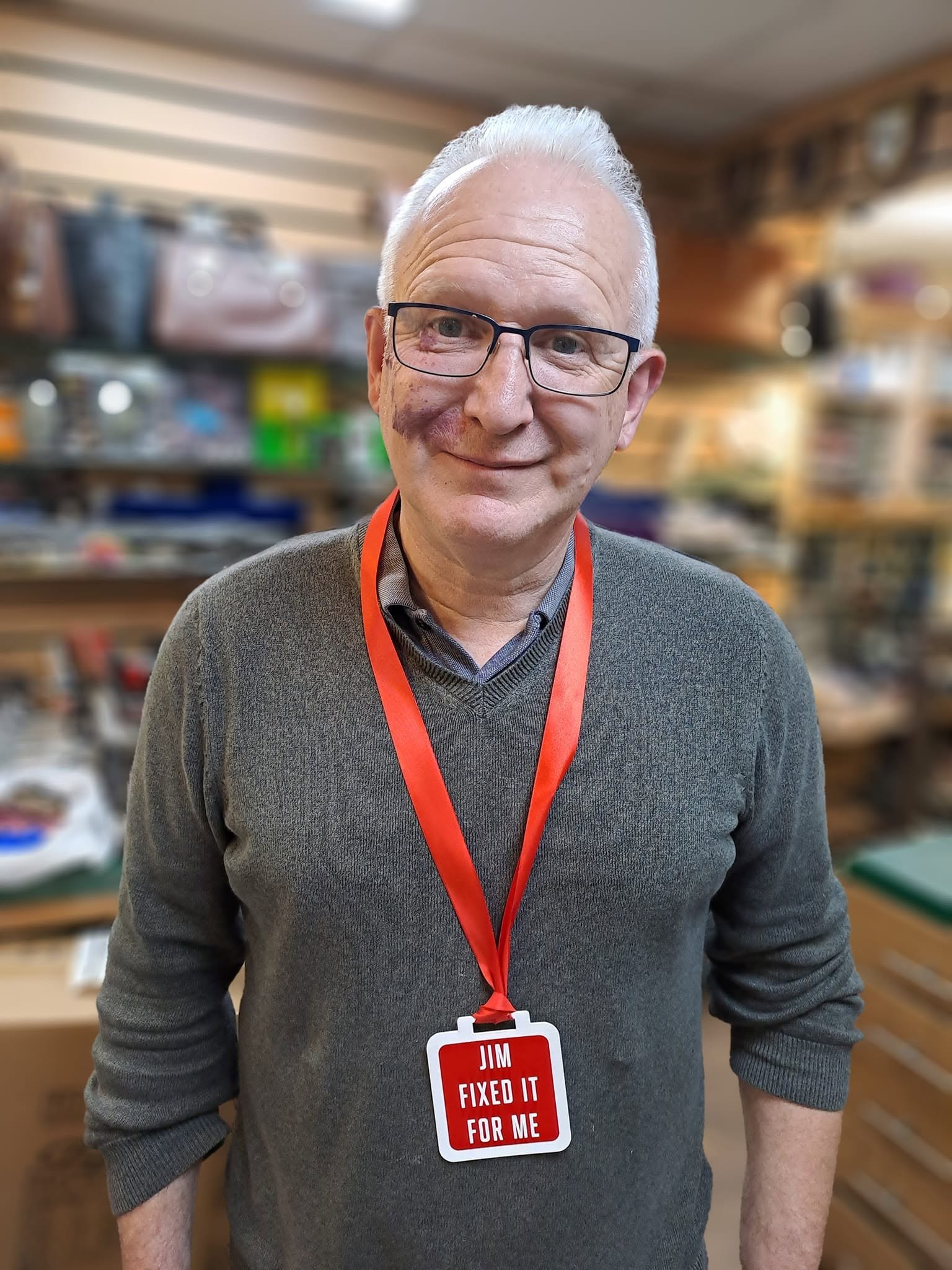 Smiling older man wearing glasses, a gray sweater, and a red name badge reading “Jim Fixed It for Me” in a workshop.