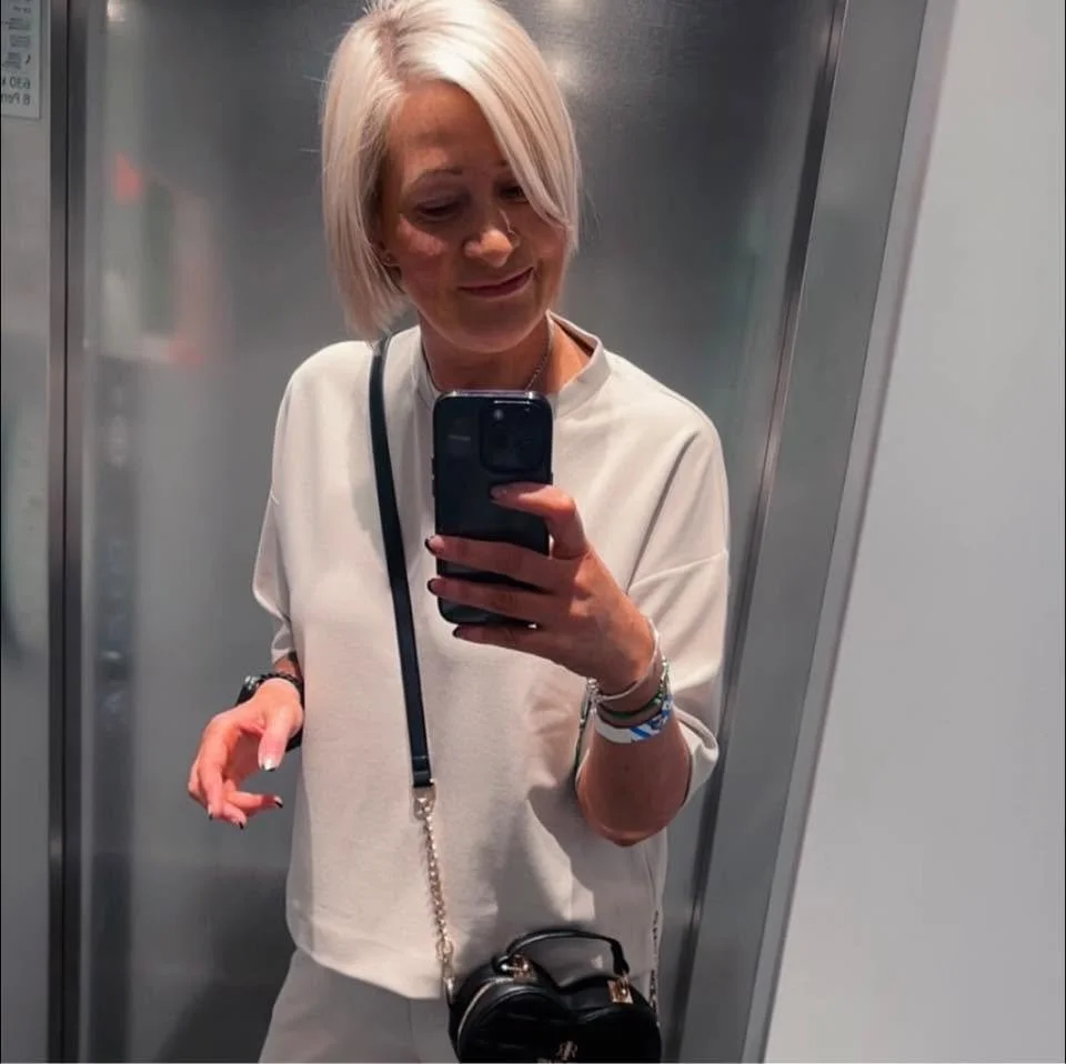 Woman with short blonde hair taking a mirror selfie in an elevator while holding a smartphone and wearing a crossbody bag.