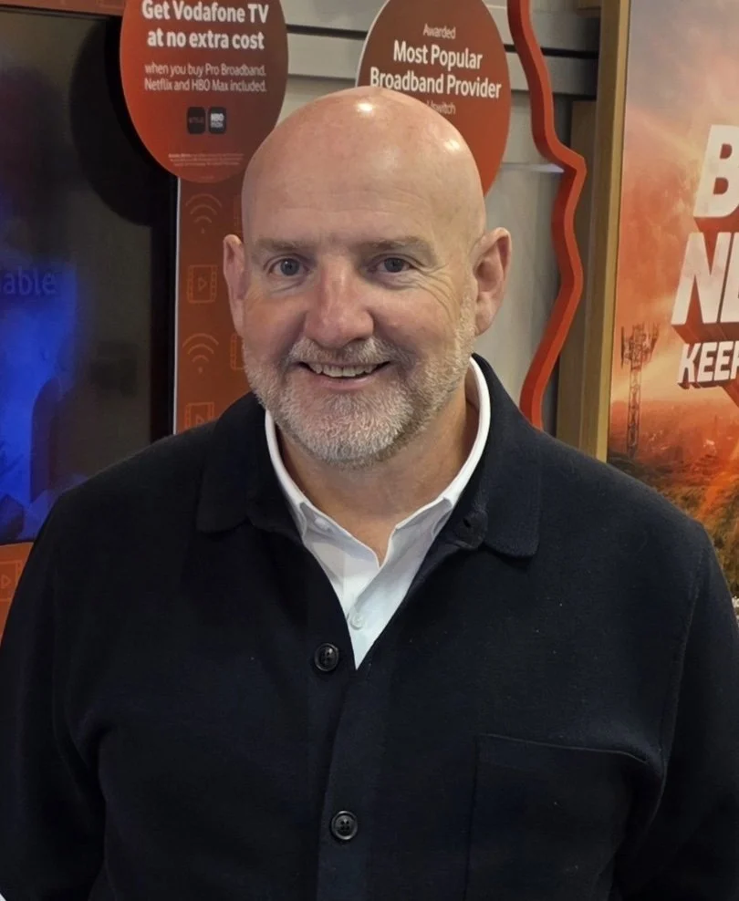 Smiling bald man in a black jacket standing in front of Vodafone broadband advertising signs