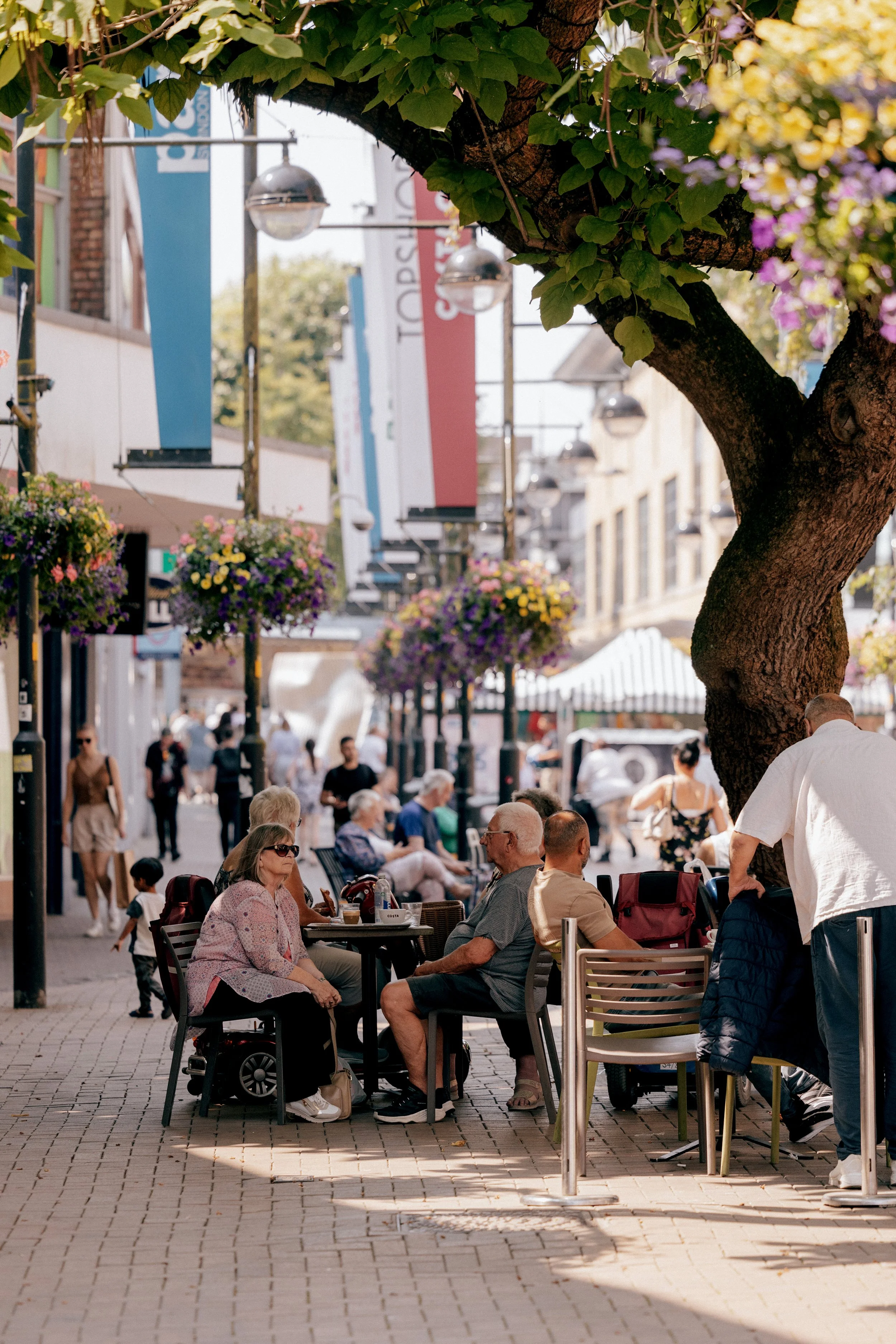 Bustling pedestrian shopping street with outdoor café seating, colorful flower baskets, hanging street lamps, and a leafy tree in the foreground