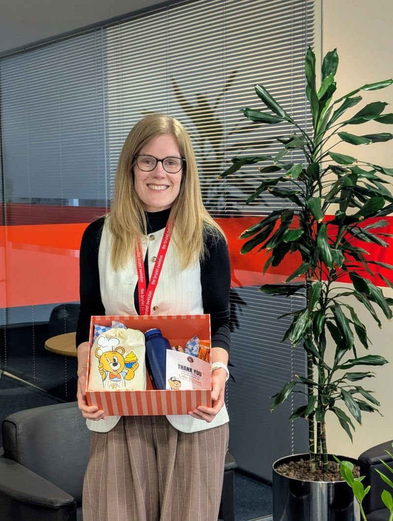 Smiling woman holding a gift box with colorful items beside a tall potted plant in an office