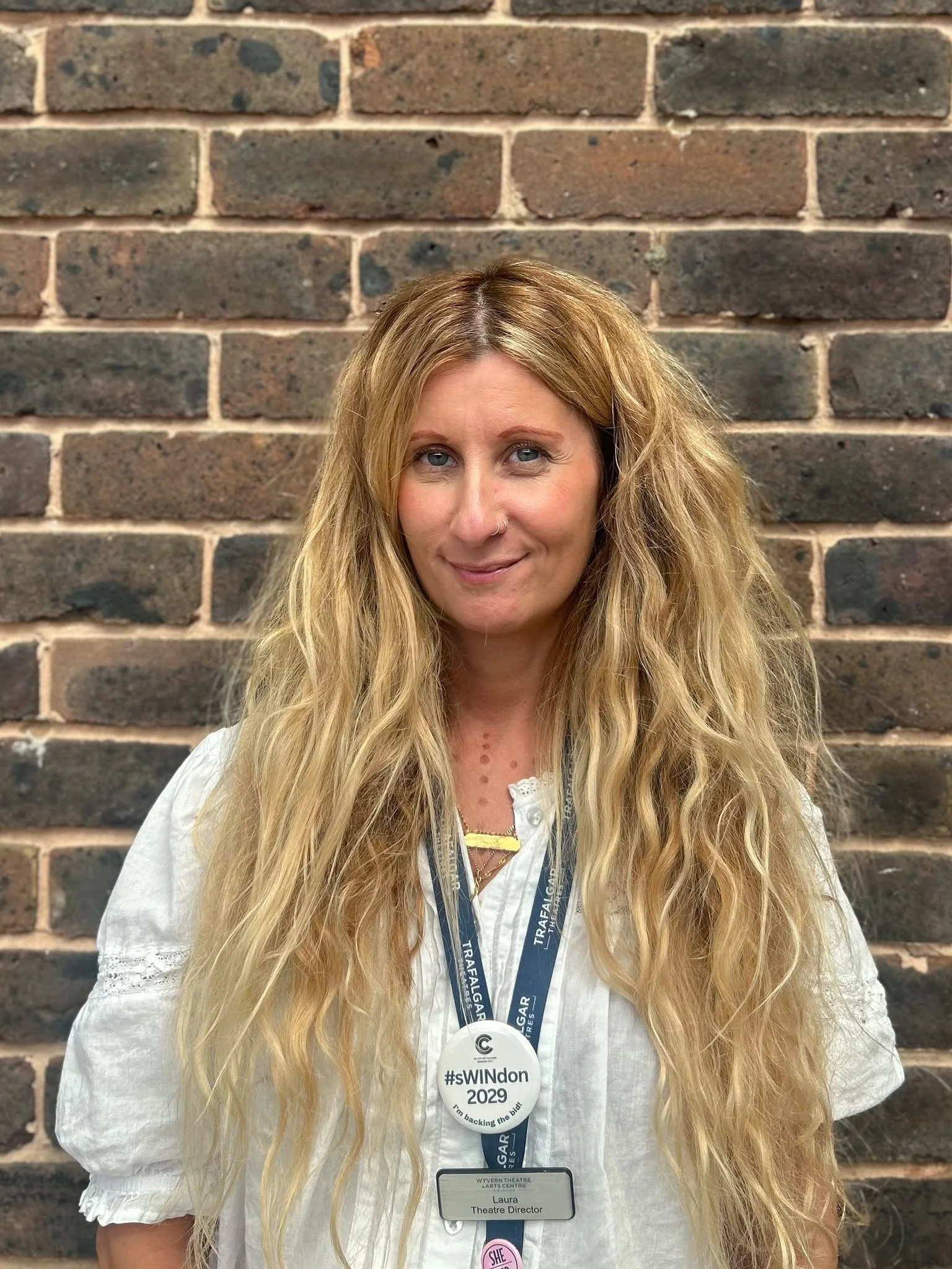Portrait of a smiling woman with long blonde hair wearing a white blouse, event lanyard, and theater director name badge in front of a brick wall.