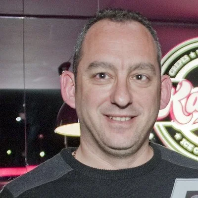 Smiling man posing indoors in front of a brightly lit restaurant sign