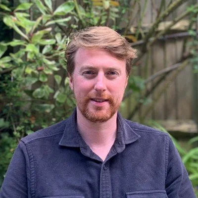 Portrait of a bearded man in a dark shirt standing outdoors in front of leafy greenery.