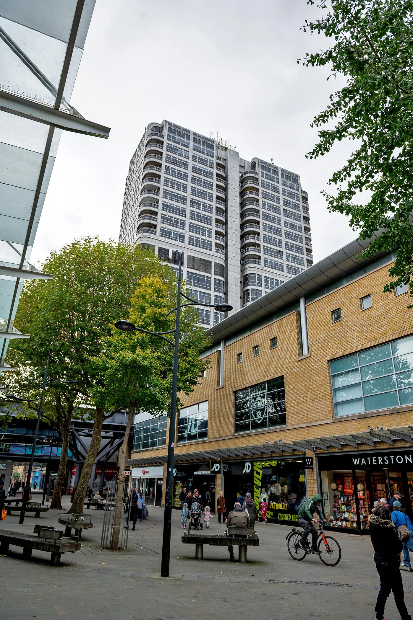 Busy pedestrian shopping plaza with Waterstones, JD Sports, trees, benches, cyclists, and a modern high-rise building. Swindon Town Centre