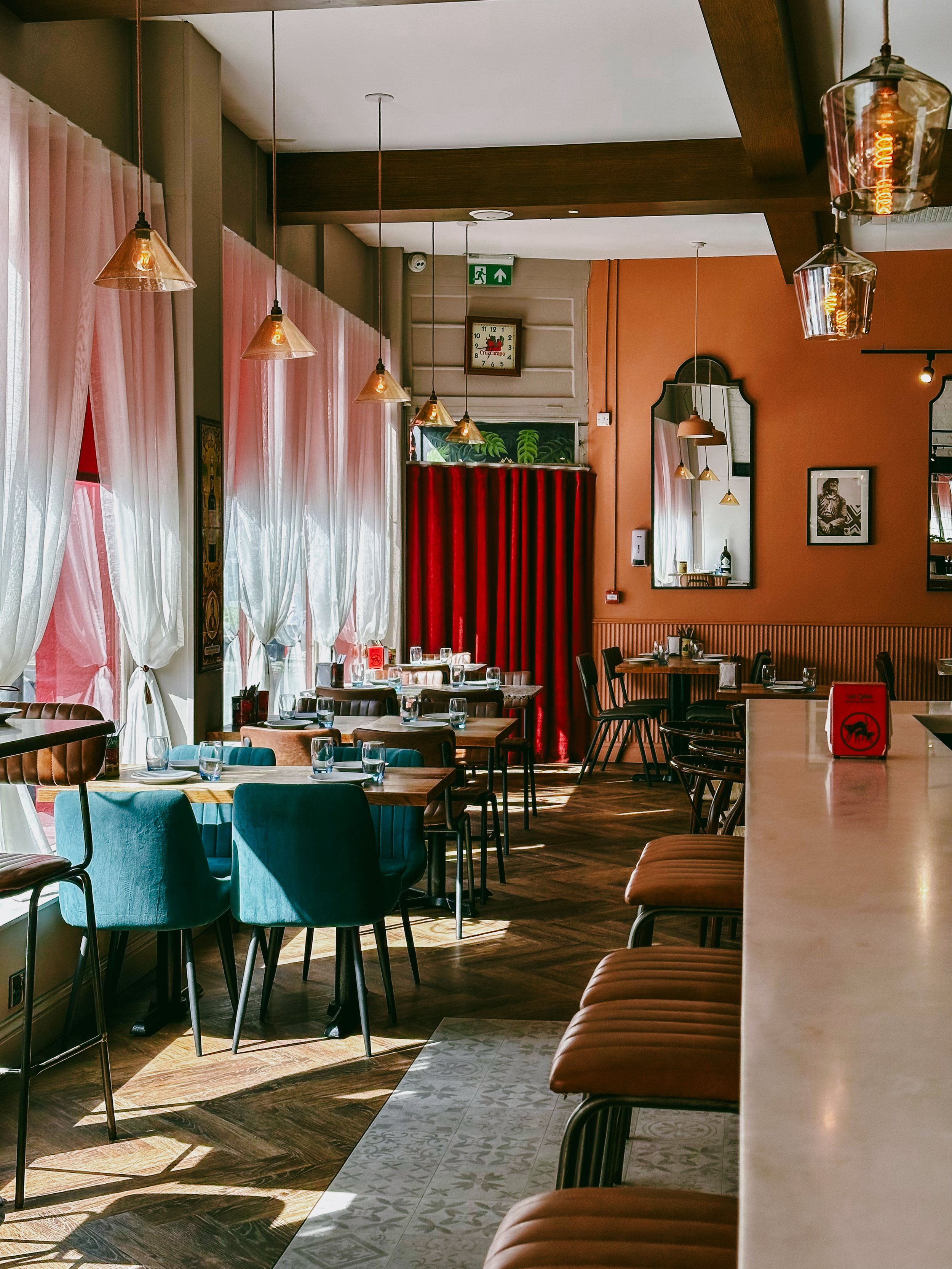 Warmly lit restaurant interior with pendant lights, pink curtains, red velvet accents, wood tables, and teal upholstered chairs