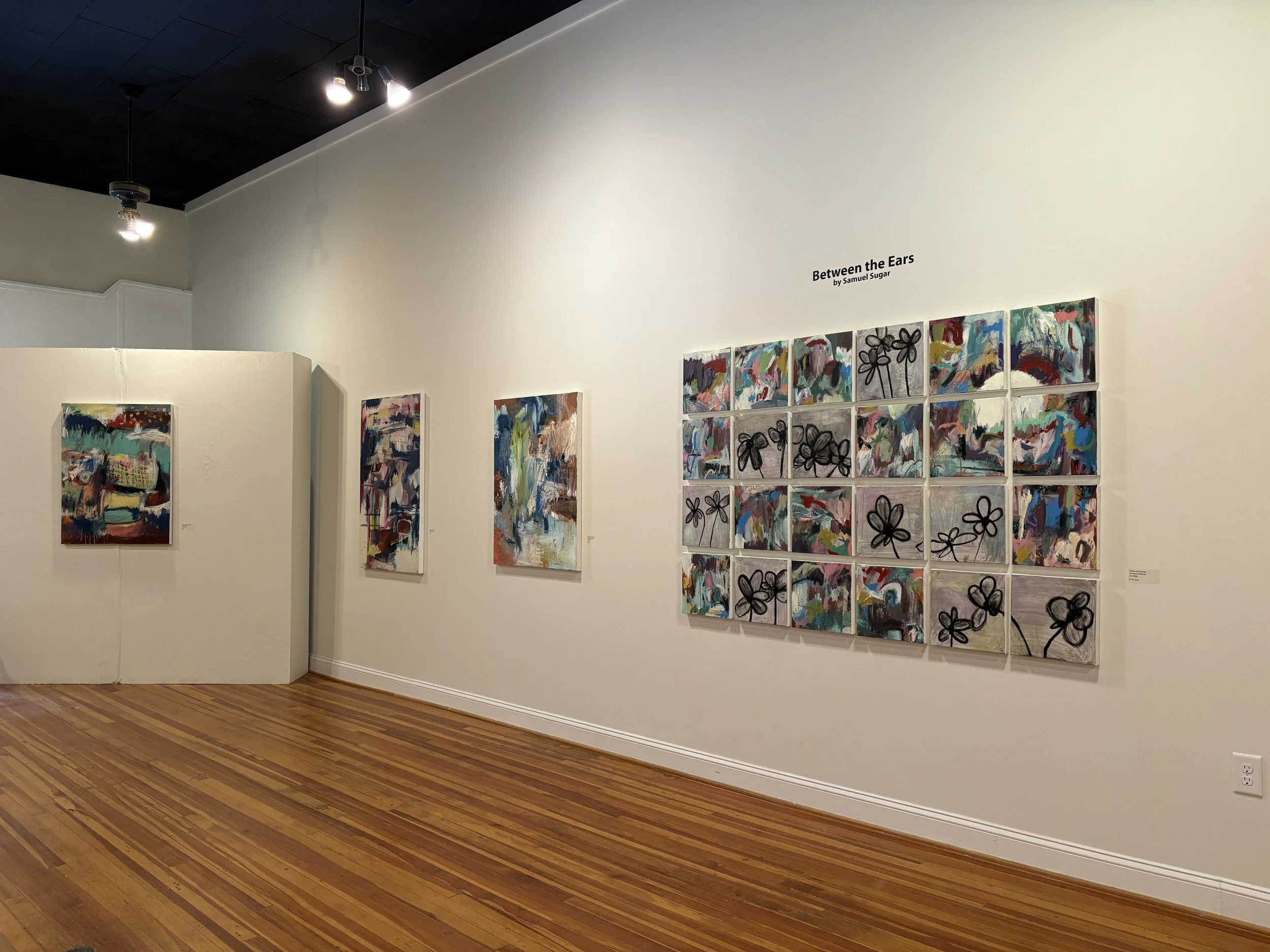 Art gallery wall displaying colorful abstract paintings, including a large floral collage titled “Between the Ears” by Samuel Sugar.