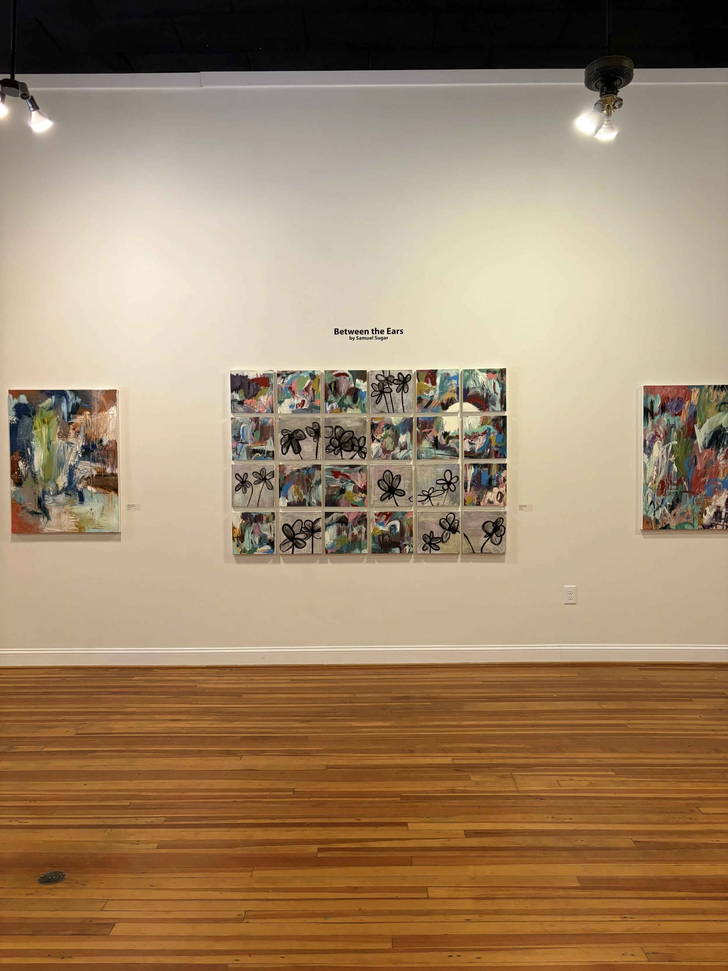 Gallery wall displaying Samuel Sugar’s abstract art exhibition “Between the Ears,” with colorful paintings and floral motifs under track lighting.