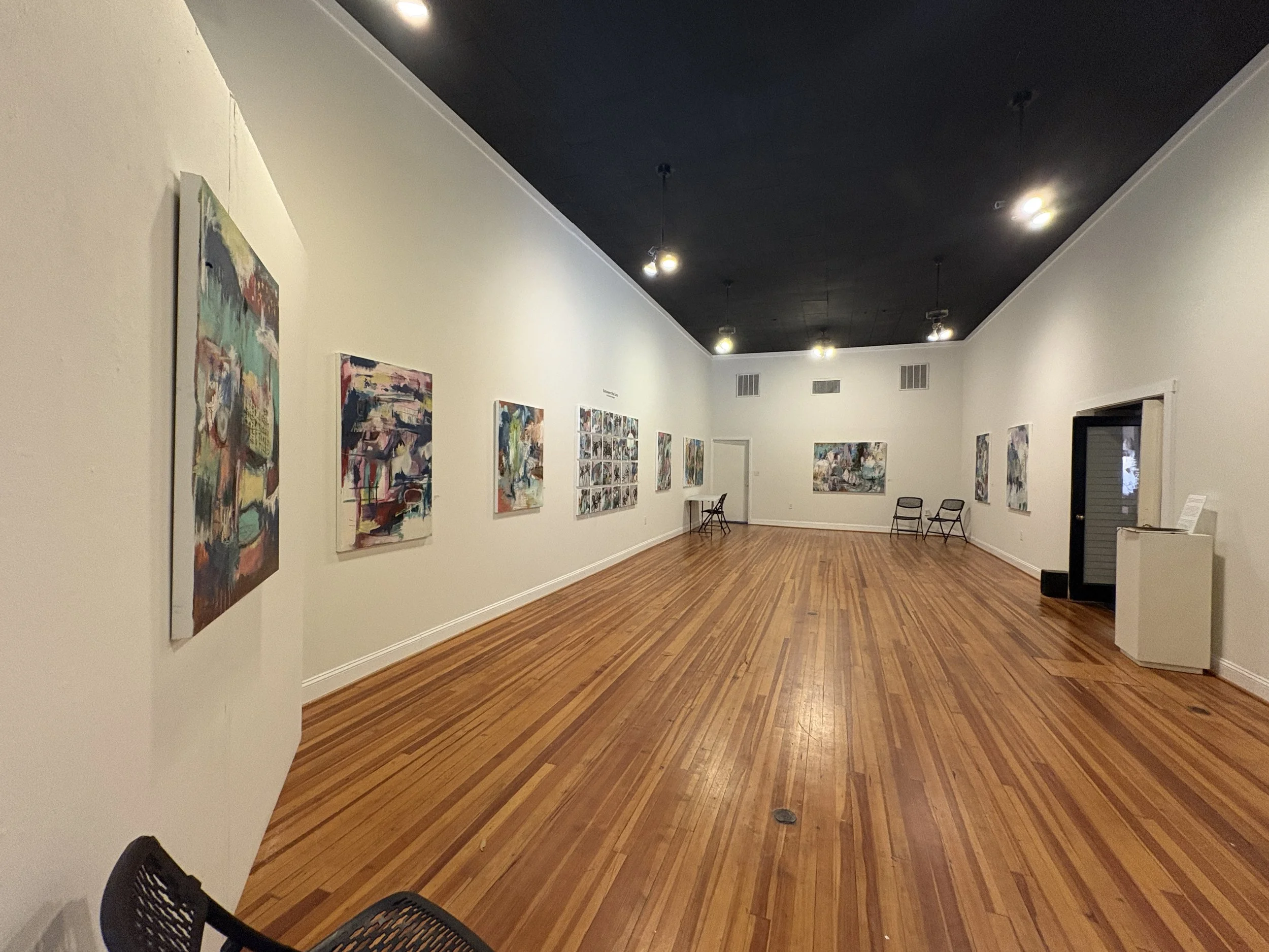 Interior of an art gallery with colorful paintings, hardwood floors, track lighting, and folding chairs