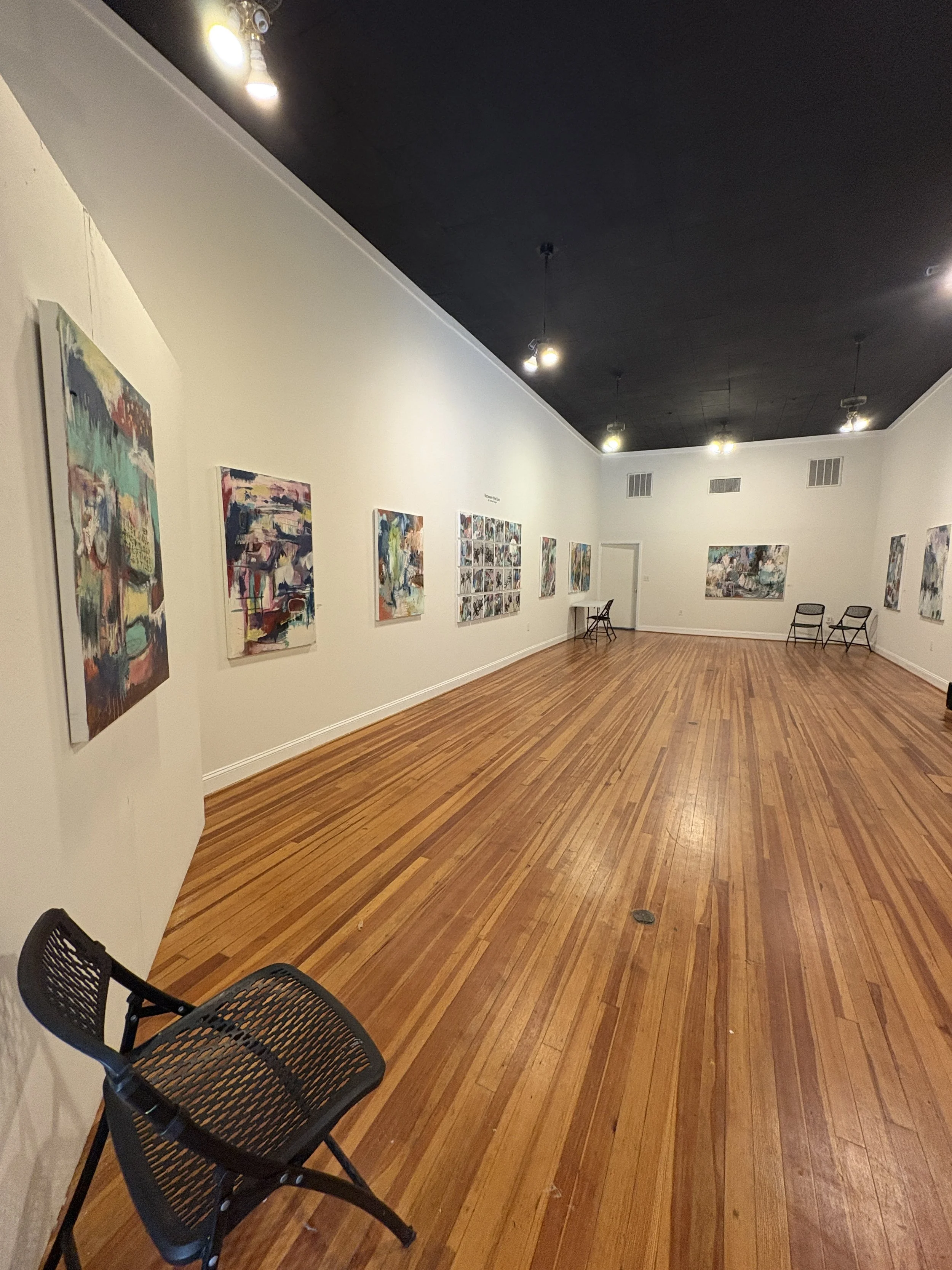 Interior of an art gallery with colorful paintings, track lighting, hardwood floors, and folding chairs.