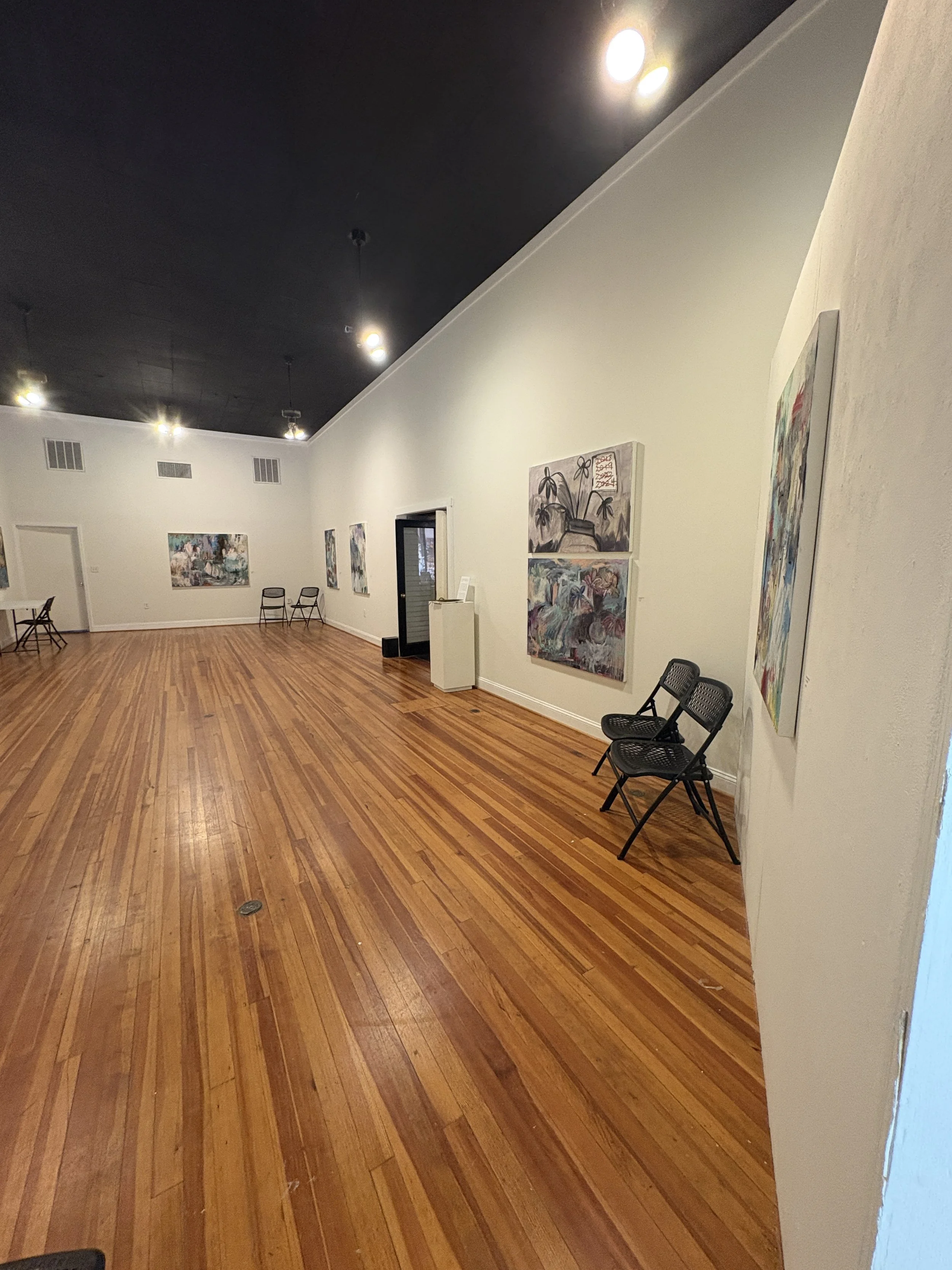 Spacious art gallery with hardwood floors, colorful paintings, folding chairs, white walls, and a black ceiling with track lighting