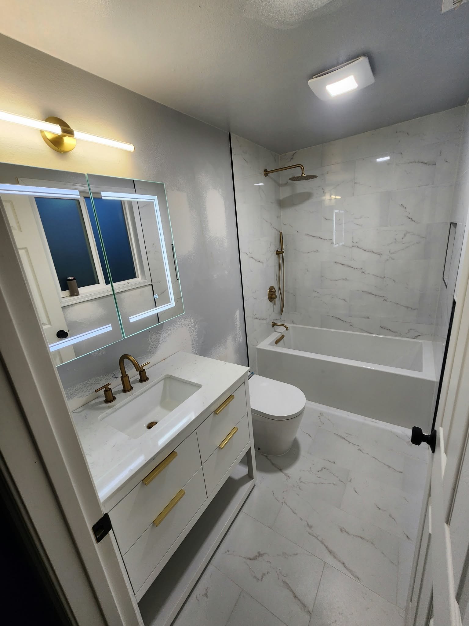 Modern white bathroom with marble-look tile, freestanding vanity, wall mirror, toilet, and bathtub shower with brushed gold fixtures.