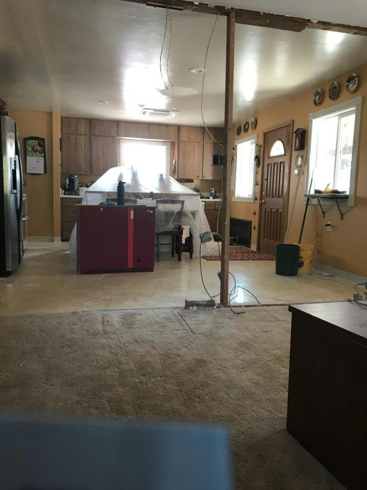 Open-plan kitchen and living area under renovation, with exposed framing, hanging electrical wires, covered furniture, and wood cabinets.