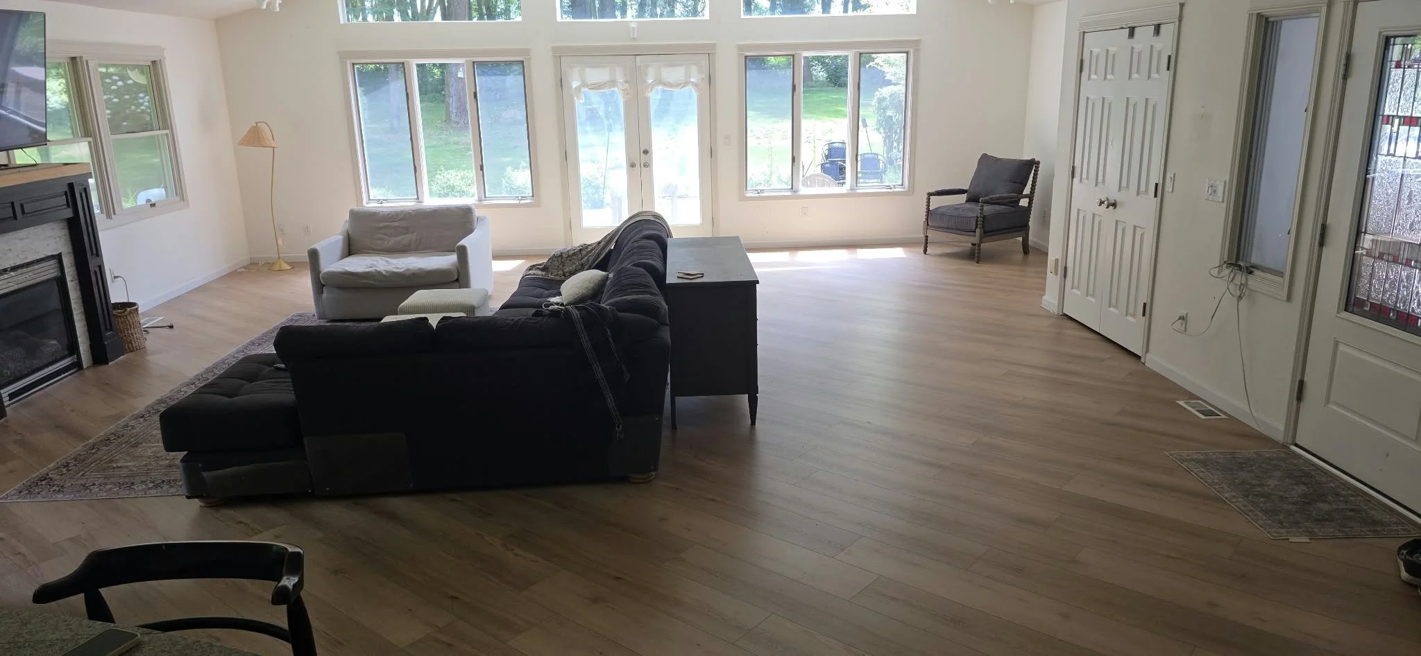 Bright open-concept living room with hardwood floors, sectional sofa, armchair, fireplace, large windows, and French doors opening to a backyard.