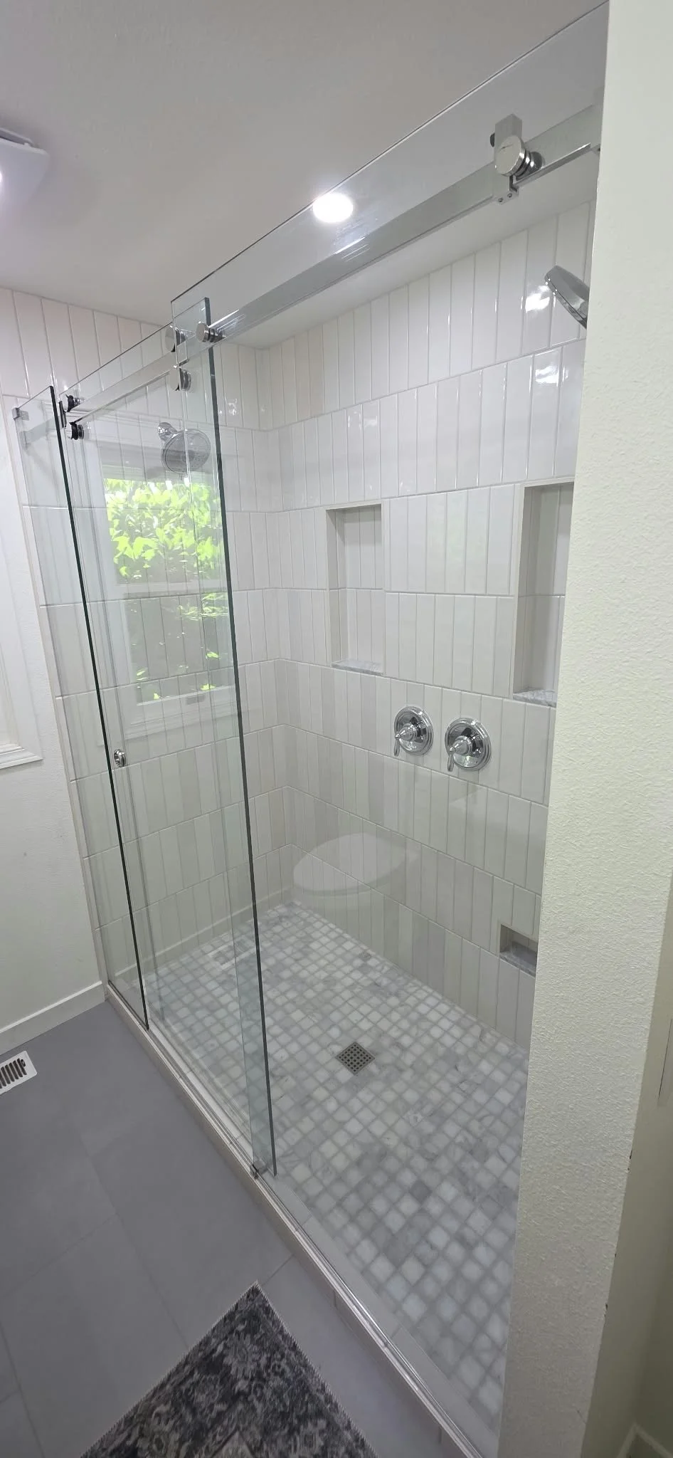 Modern walk-in shower with frameless glass enclosure, white vertical tile walls, built-in niches, chrome fixtures, and mosaic tile floor