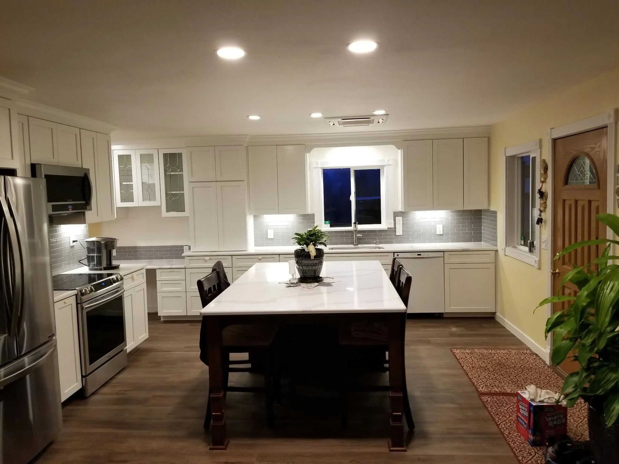 Bright modern white kitchen with a large island, stainless steel appliances, gray subway-tile backsplash, and a central sink beneath a window.