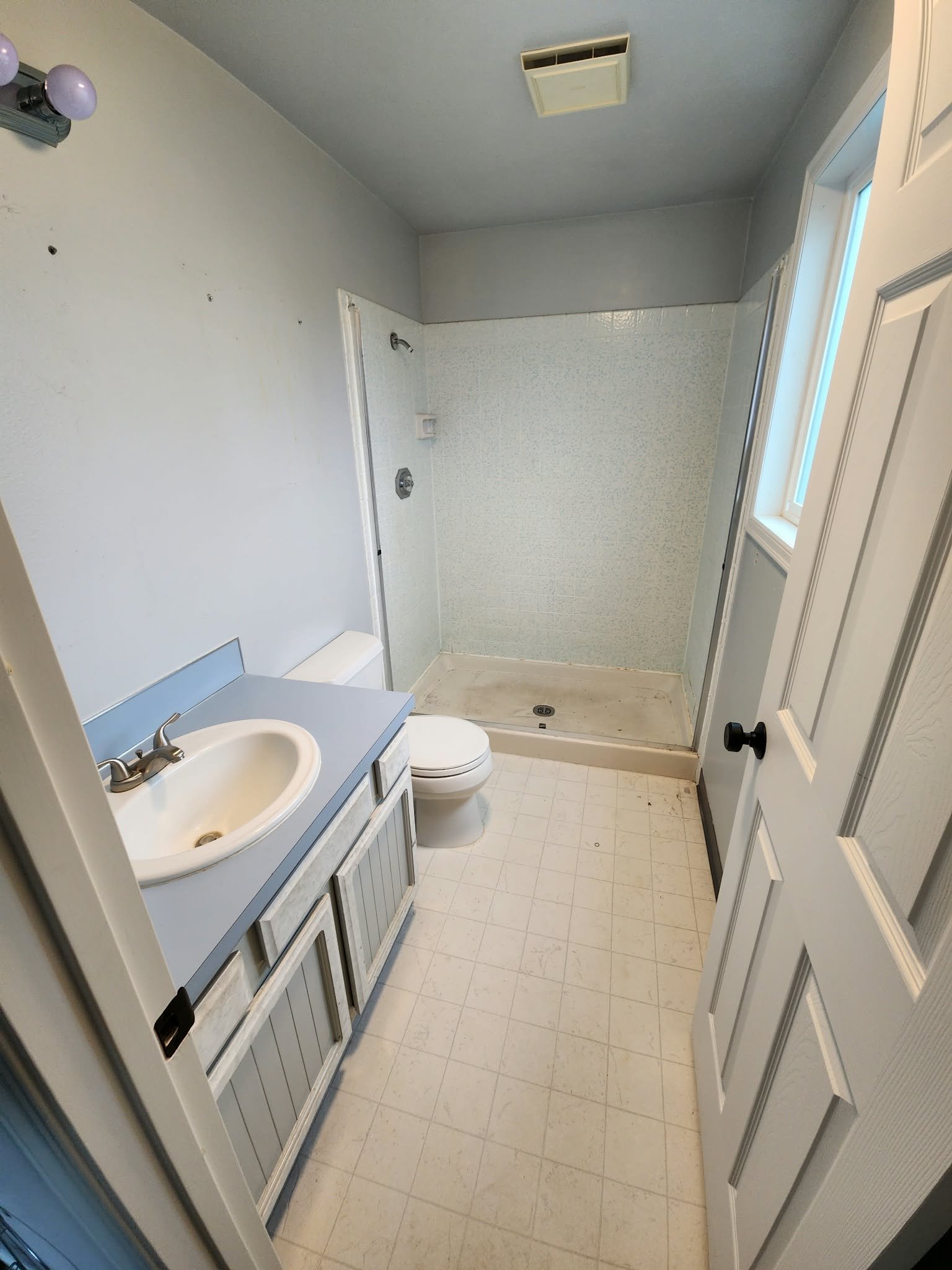 Small bathroom with blue vanity and sink, toilet, tiled shower, window, and off-white tile floor.