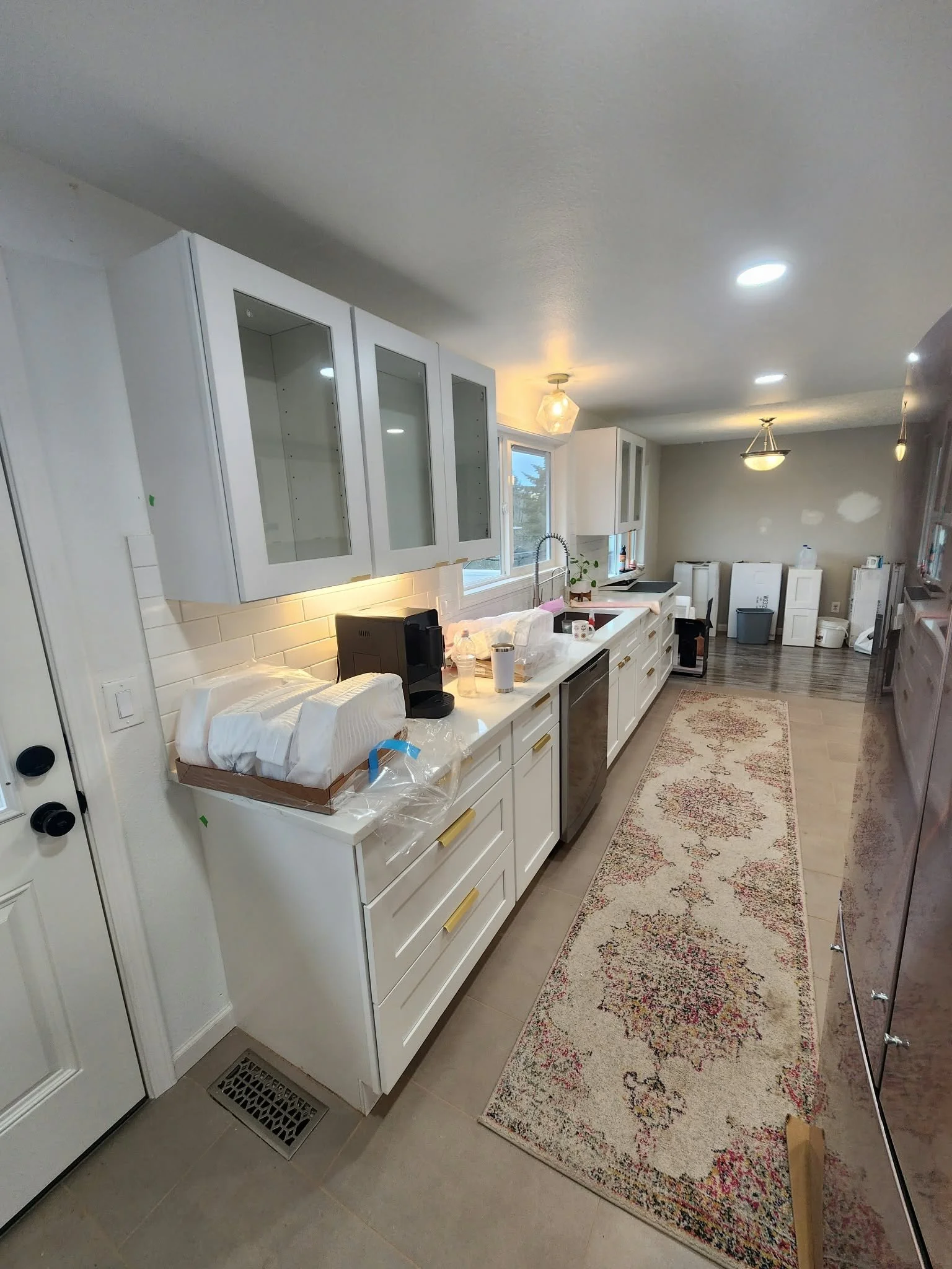 Bright modern white kitchen with glass-front cabinets, island, stainless-steel appliances, pendant lighting, and a patterned runner rug.