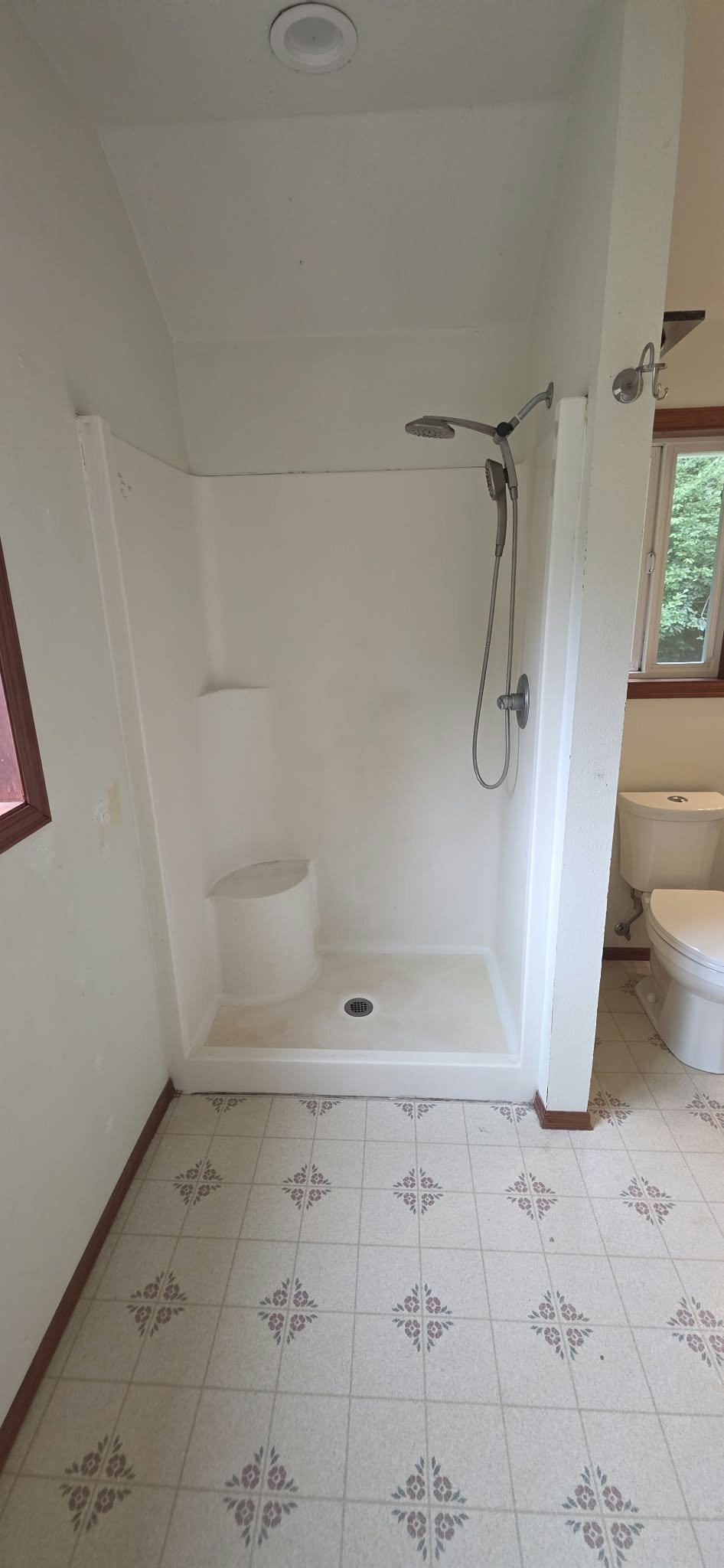 White bathroom with a walk-in shower, handheld showerhead, toilet, window, and patterned tile floor