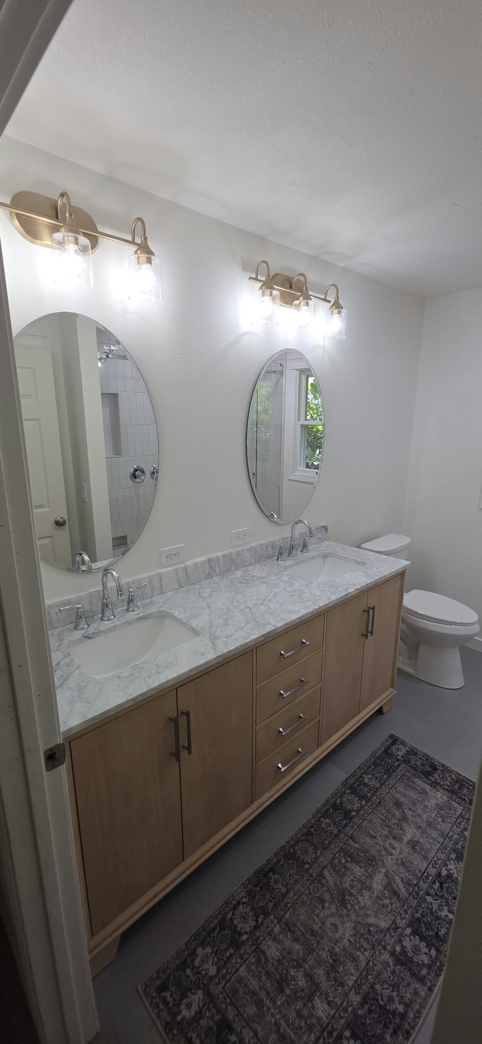 Bright bathroom with double vanity, marble countertop, oval mirrors, brass light fixtures, and toilet