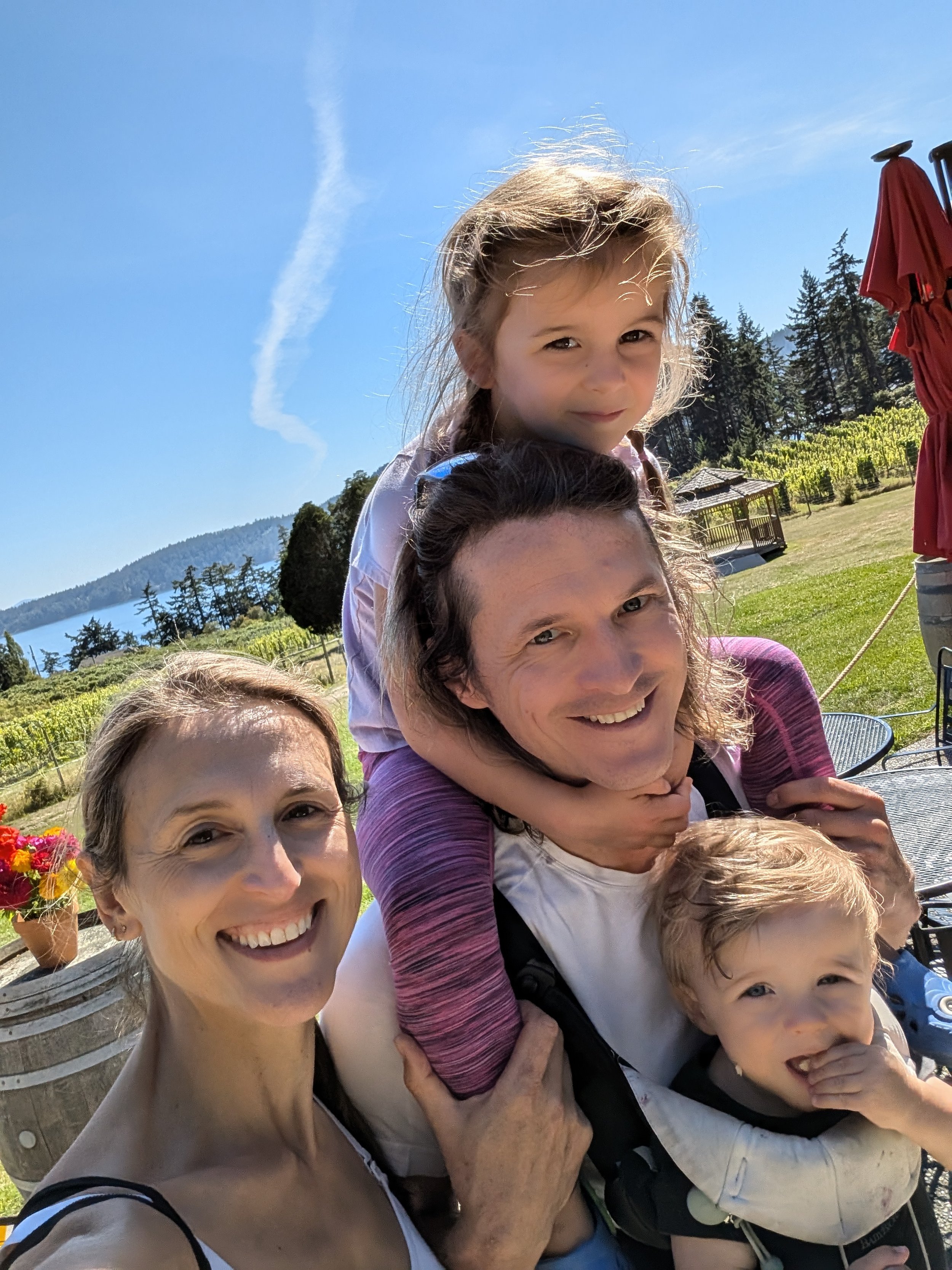 Smiling family taking a sunny outdoor selfie in a garden with mountain and lake views.