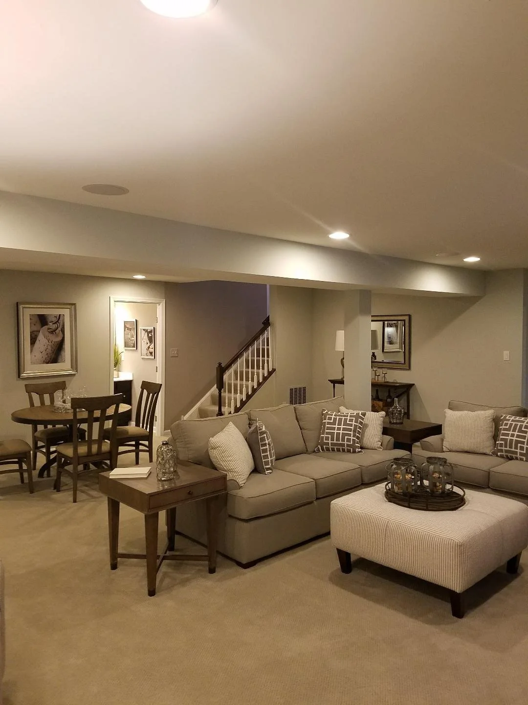 Cozy beige living room basement with sectional sofa, ottoman, coffee tables, dining area, and staircase