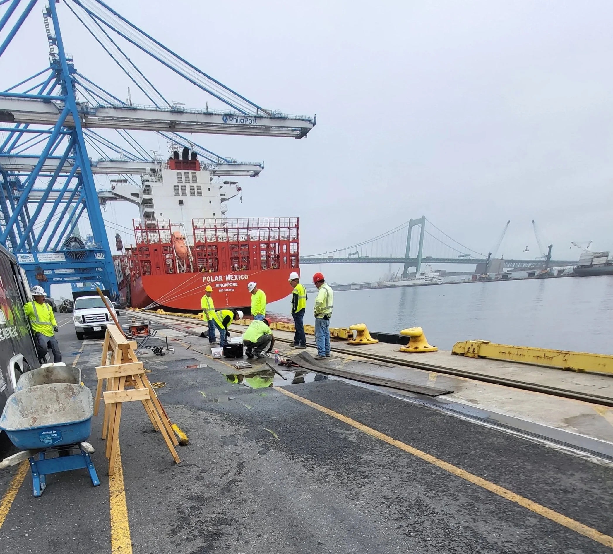 Concrete repair specialists in safety gear fixing the crane tracks with the Polar Mexico cargo ship beneath blue Philaport container cranes.