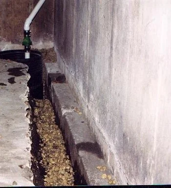 Narrow concrete drainage channel filled with leaves and debris beside a weathered wall, with a hose and pipe above