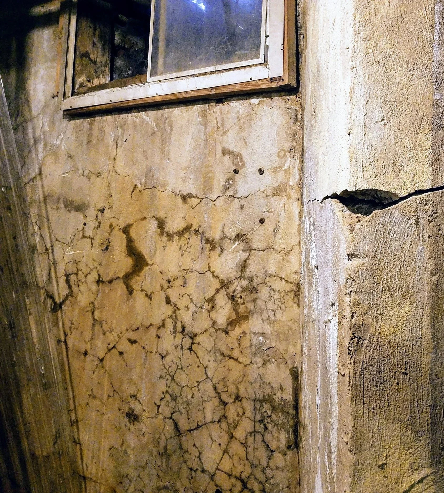 Cracked, weathered plaster wall with a small dirty window and exposed structural damage