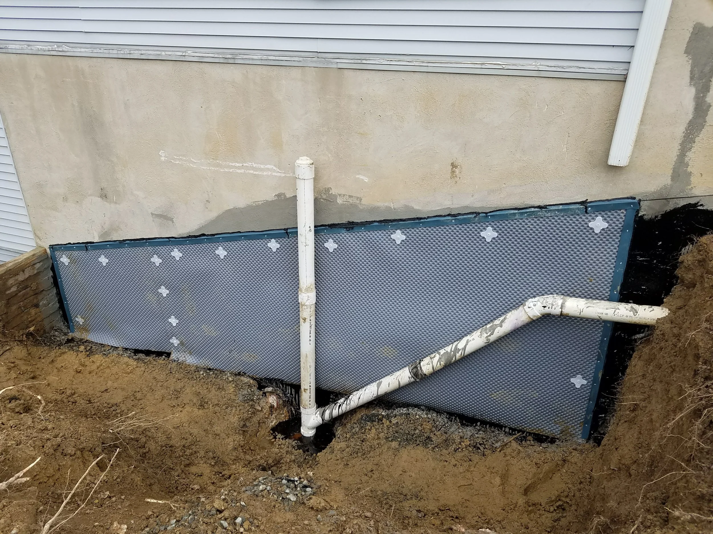Exterior foundation waterproofing with drainage membrane and PVC plumbing pipes exposed in excavated soil