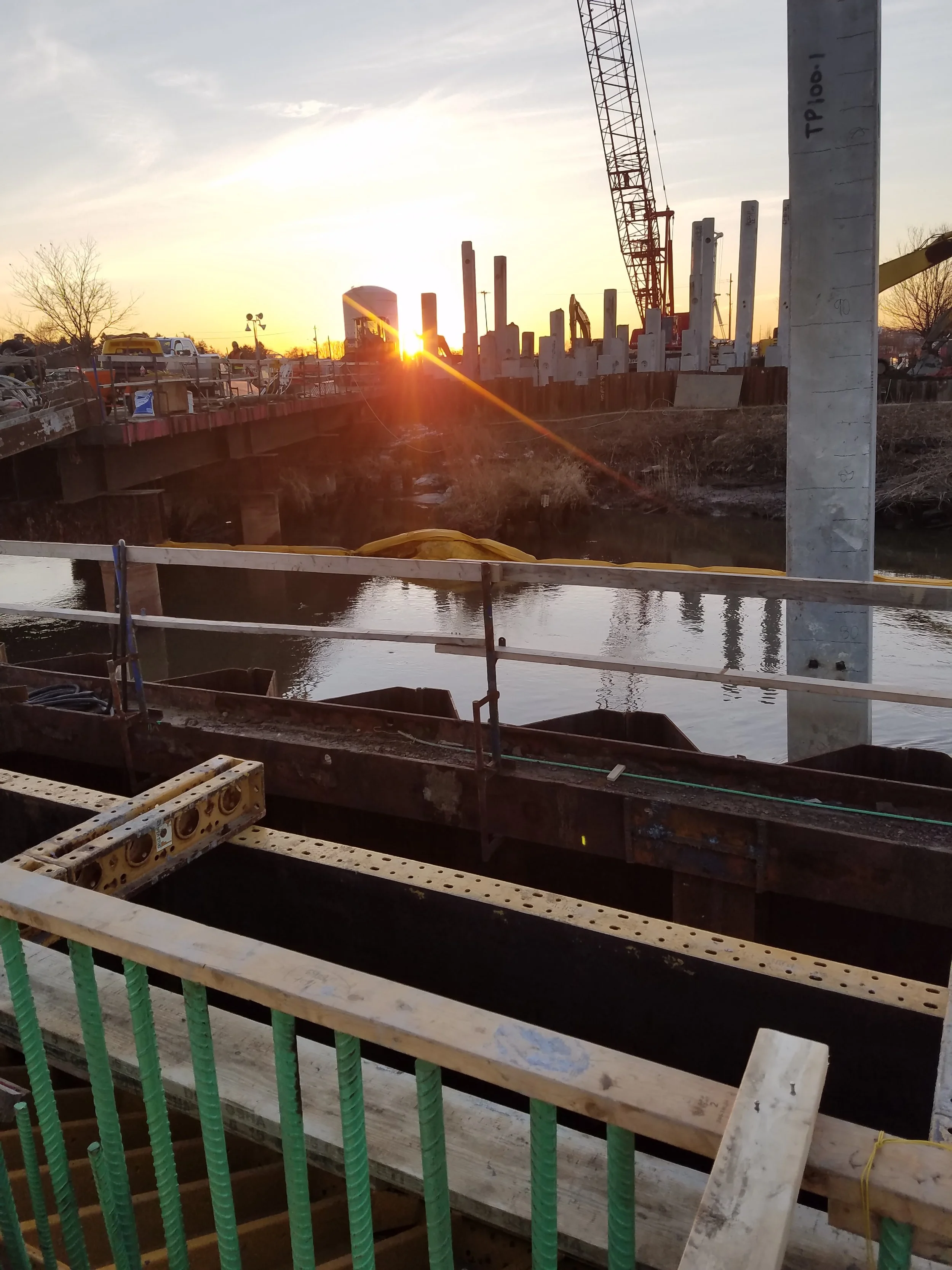 Bridge construction site at sunset with concrete columns, steel rebar, formwork, excavated foundations, water, and a tower crane.