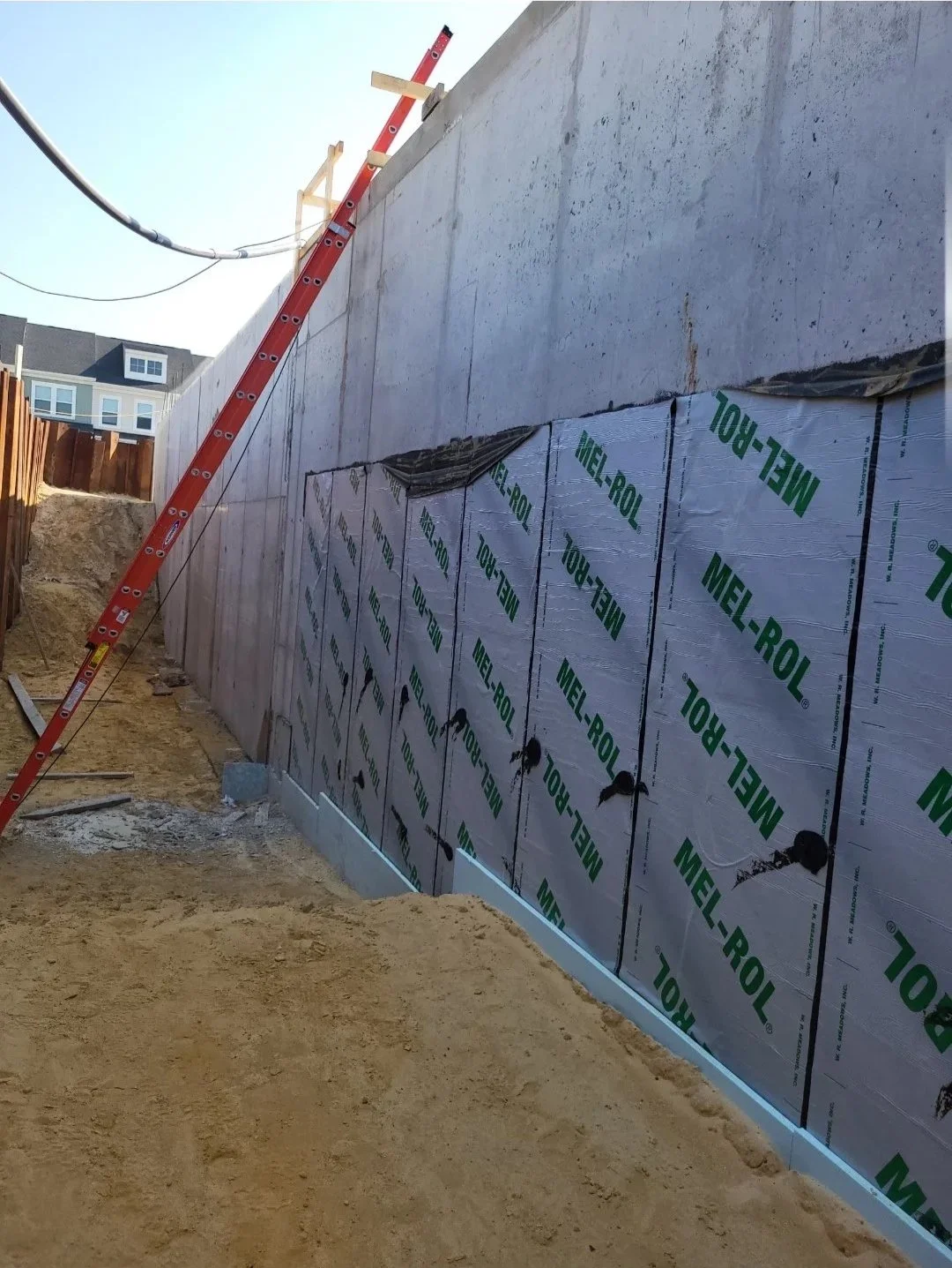 Construction site with a red extension ladder leaning against a concrete retaining wall insulated with foil-faced panels.
