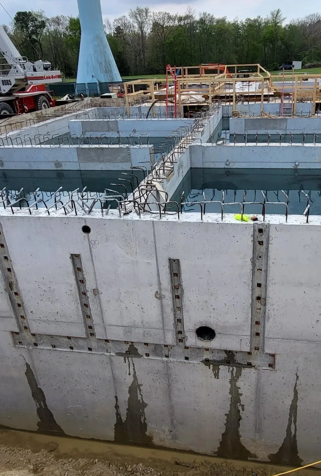 Concrete foundation walls with exposed rebar under construction, wooden forms, and a Link-Belt crane at a wooded construction site.