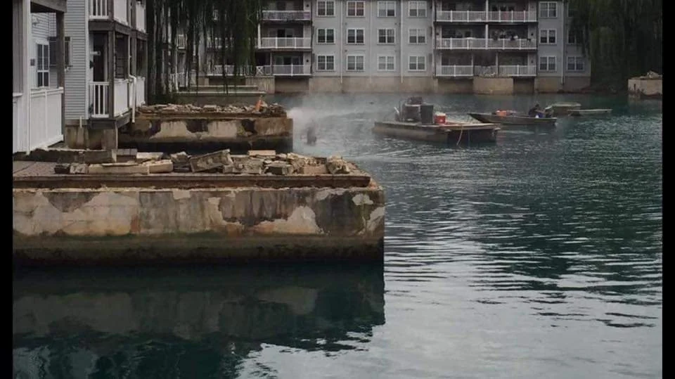 Damaged waterfront apartment buildings beside a flooded marina with small boats and smoke rising from the water
