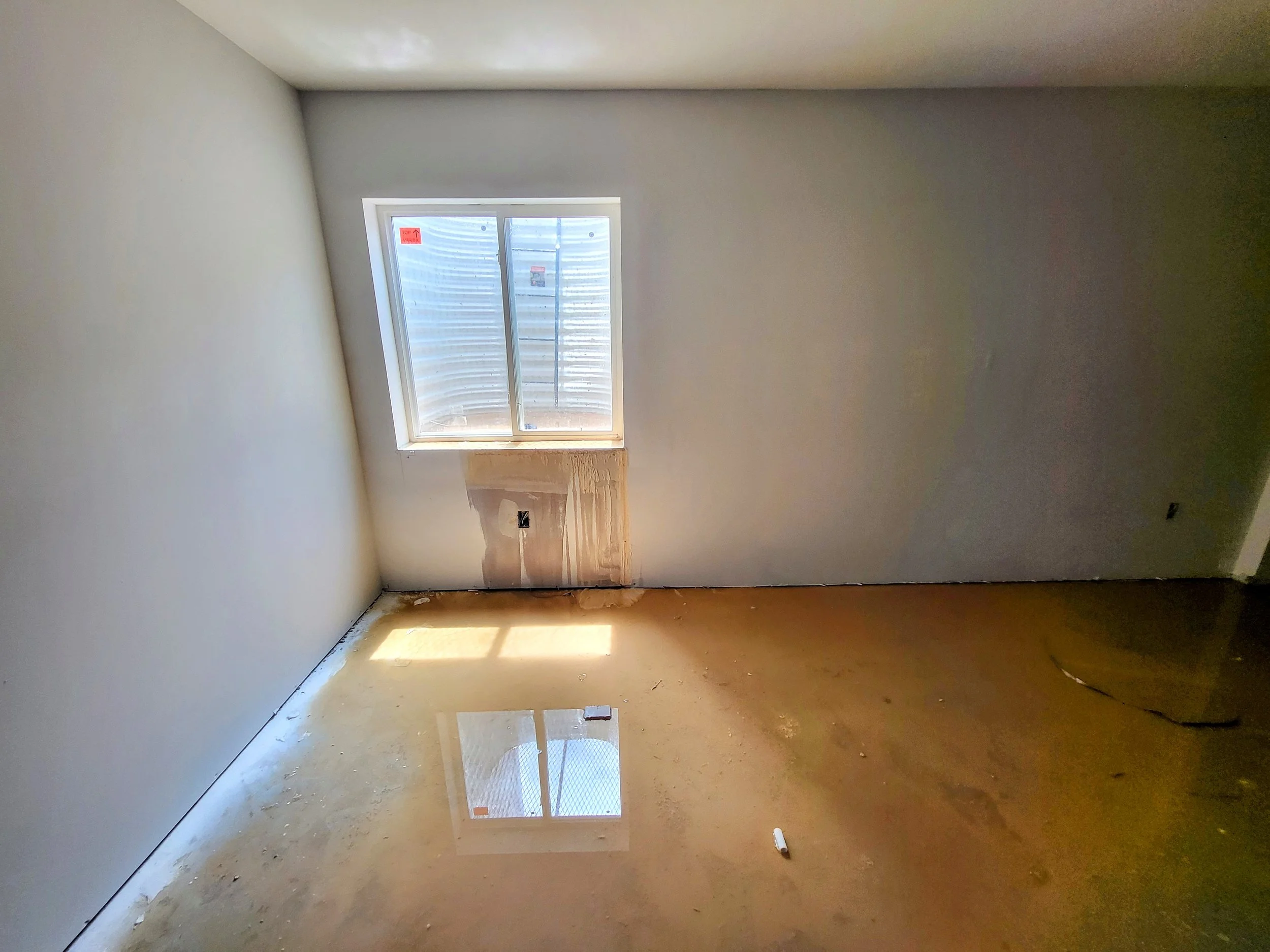 Unfinished empty room with a window, patchy walls, exposed electrical outlet, and dusty concrete floor.