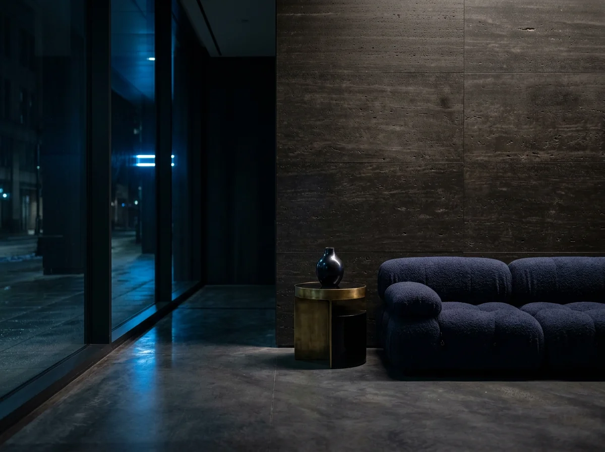 Modern dark lounge with a navy blue sofa, black vase on a brass side table, concrete wall, and blue-lit glass corridor