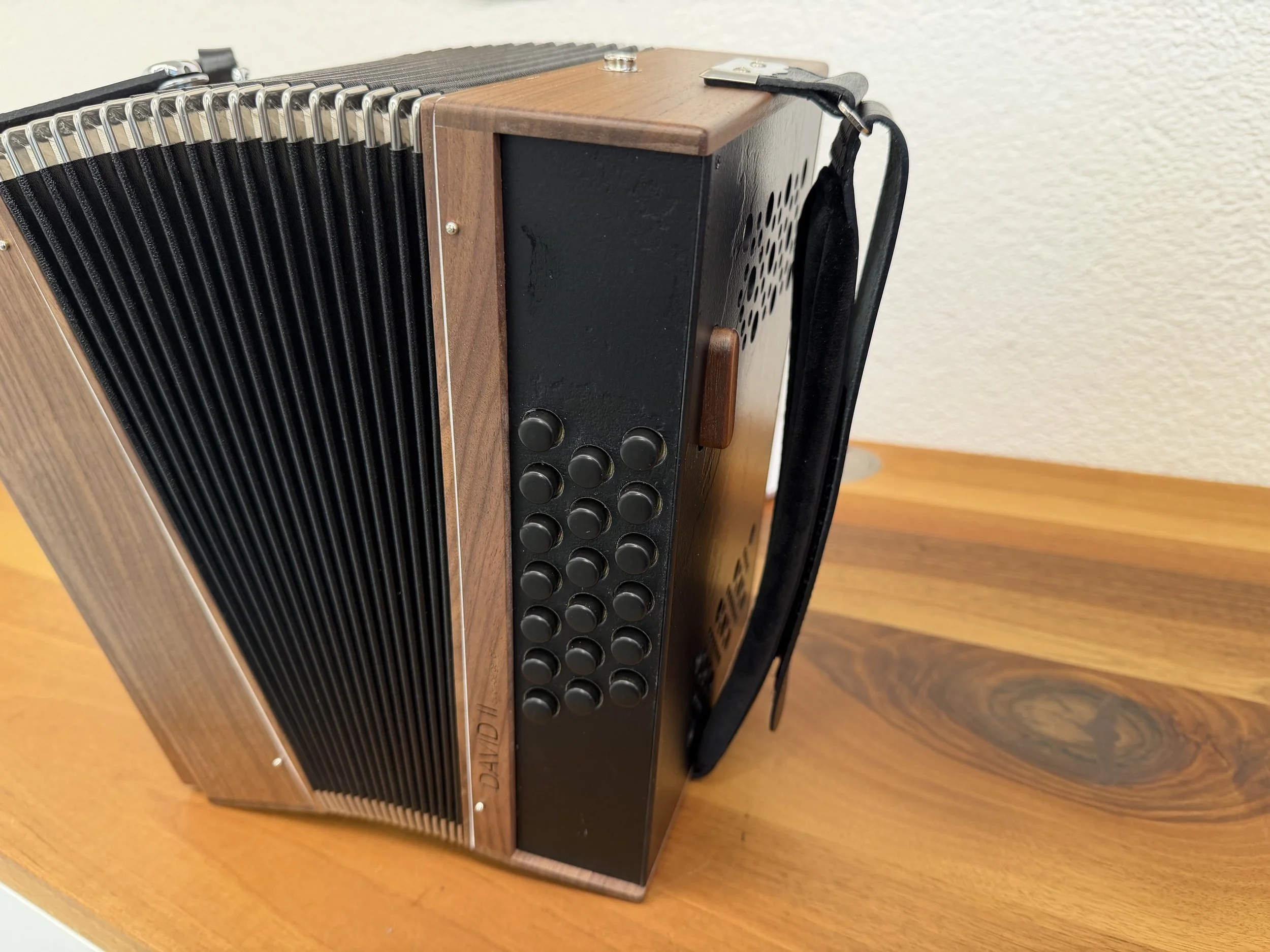 Nahaufnahme eines hölzernen Akkordeons mit schwarzen Tasten, Metallknöpfen und schwarzem Trageriemen auf einem Holztisch