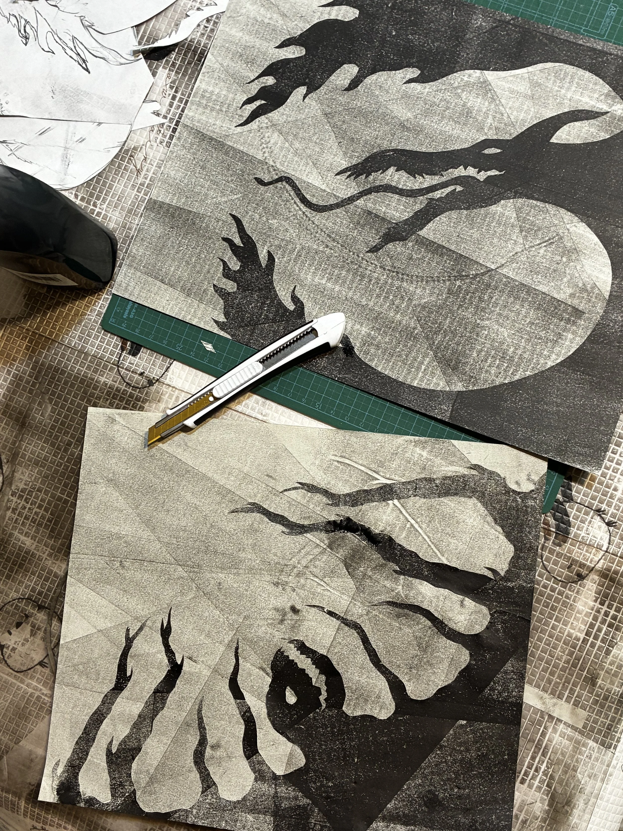 Black-and-white linocut print of abstract animal silhouettes on a cutting mat, surrounded by tracing paper and a utility knife