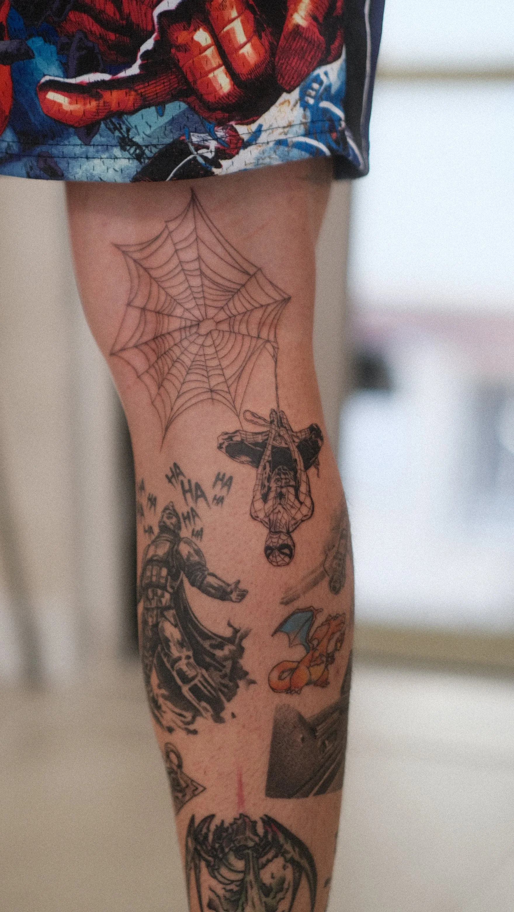 Gros plan sur un bras couvert de tatouages colorés et en noir et blanc, avec une toile d’araignée, des illustrations de bande dessinée et un short à motif Spider-Man.