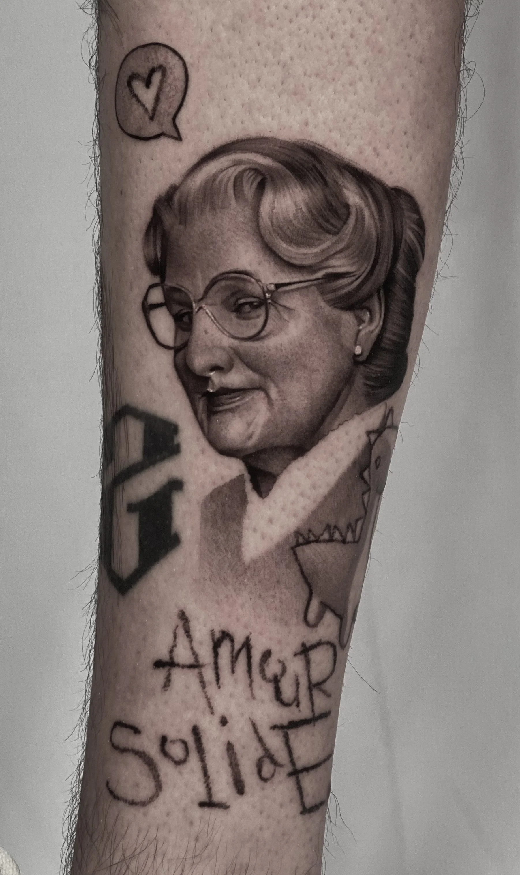 Gros plan d’un tatouage en noir et gris représentant une femme aux lunettes, avec un cœur, des motifs floraux et l’inscription « Amour toujours » sur un bras.
