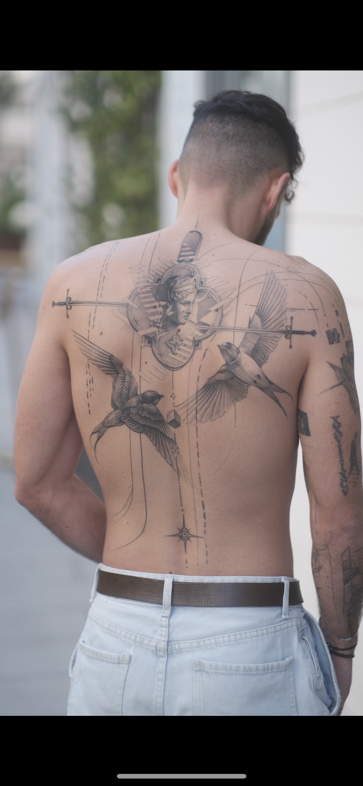Homme torse nu vu de dos, portant un grand tatouage artistique représentant des oiseaux, une figure centrale et des épées.